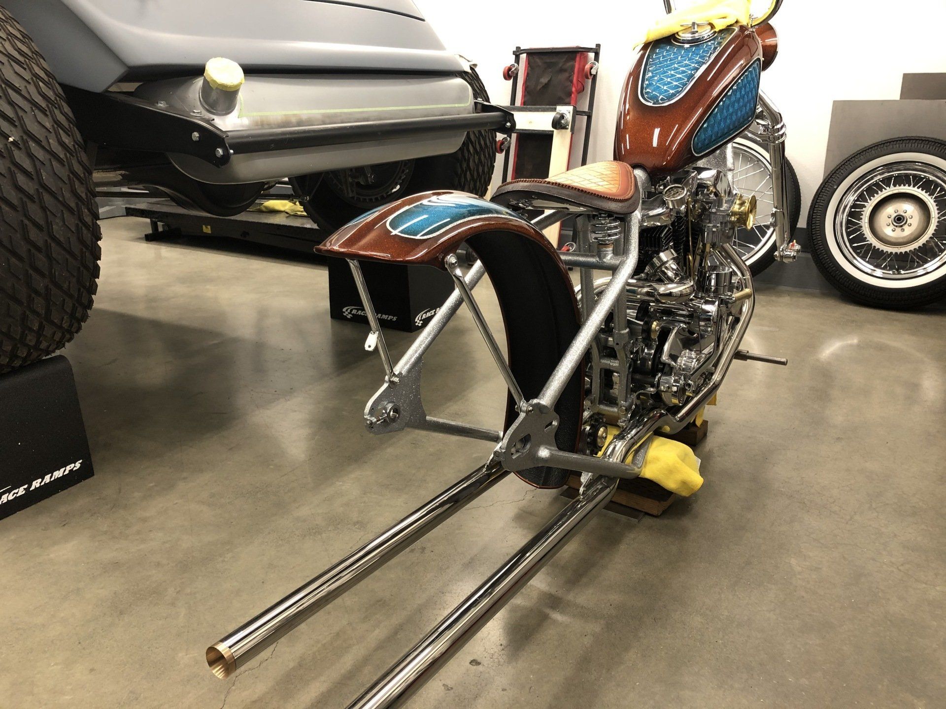 Custom motorcycle with blue and orange paint, chrome details, and long front fork on display indoors.