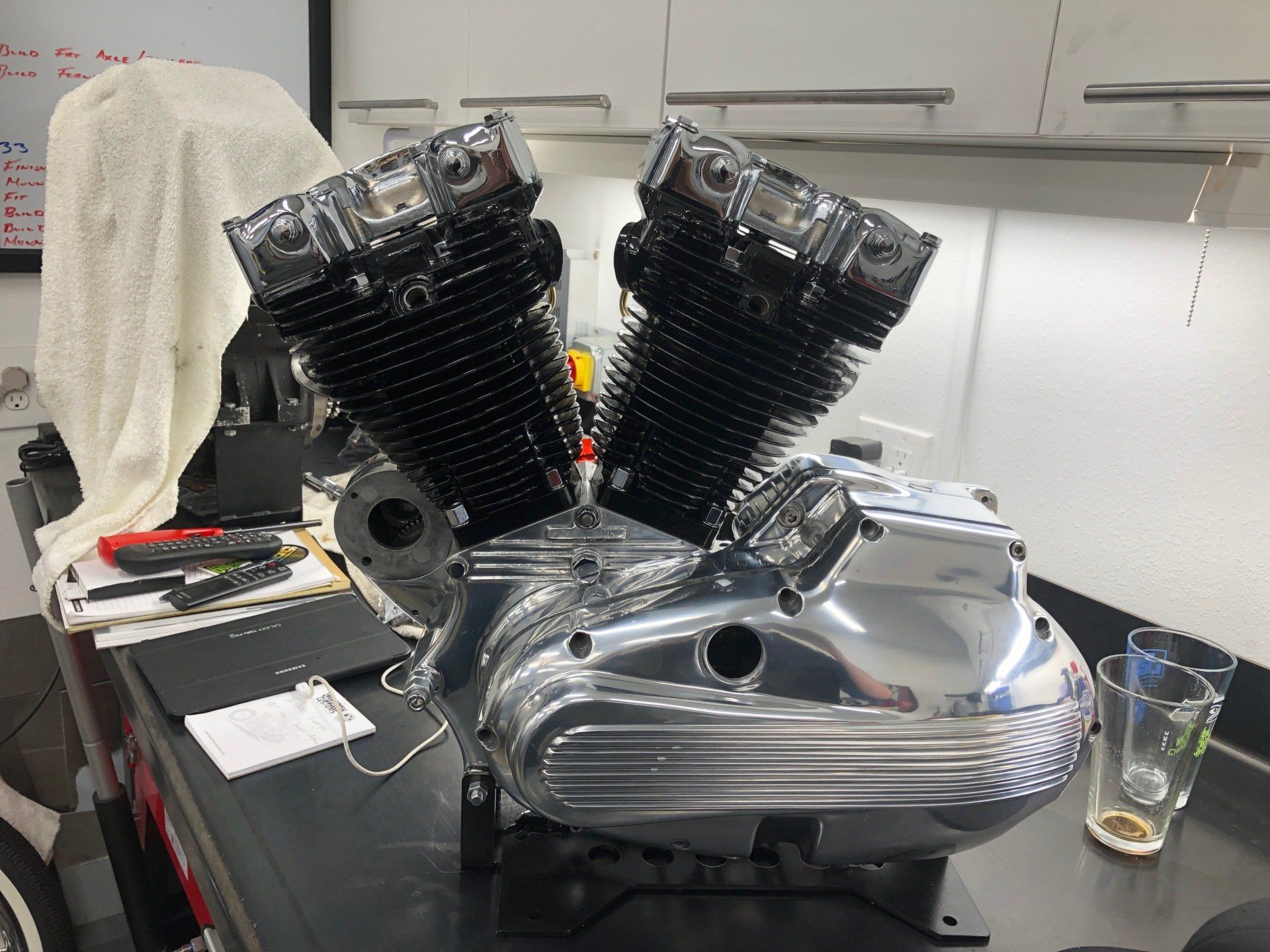 Chrome V-twin motorcycle engine on a workbench in a garage, with a drink glass nearby.