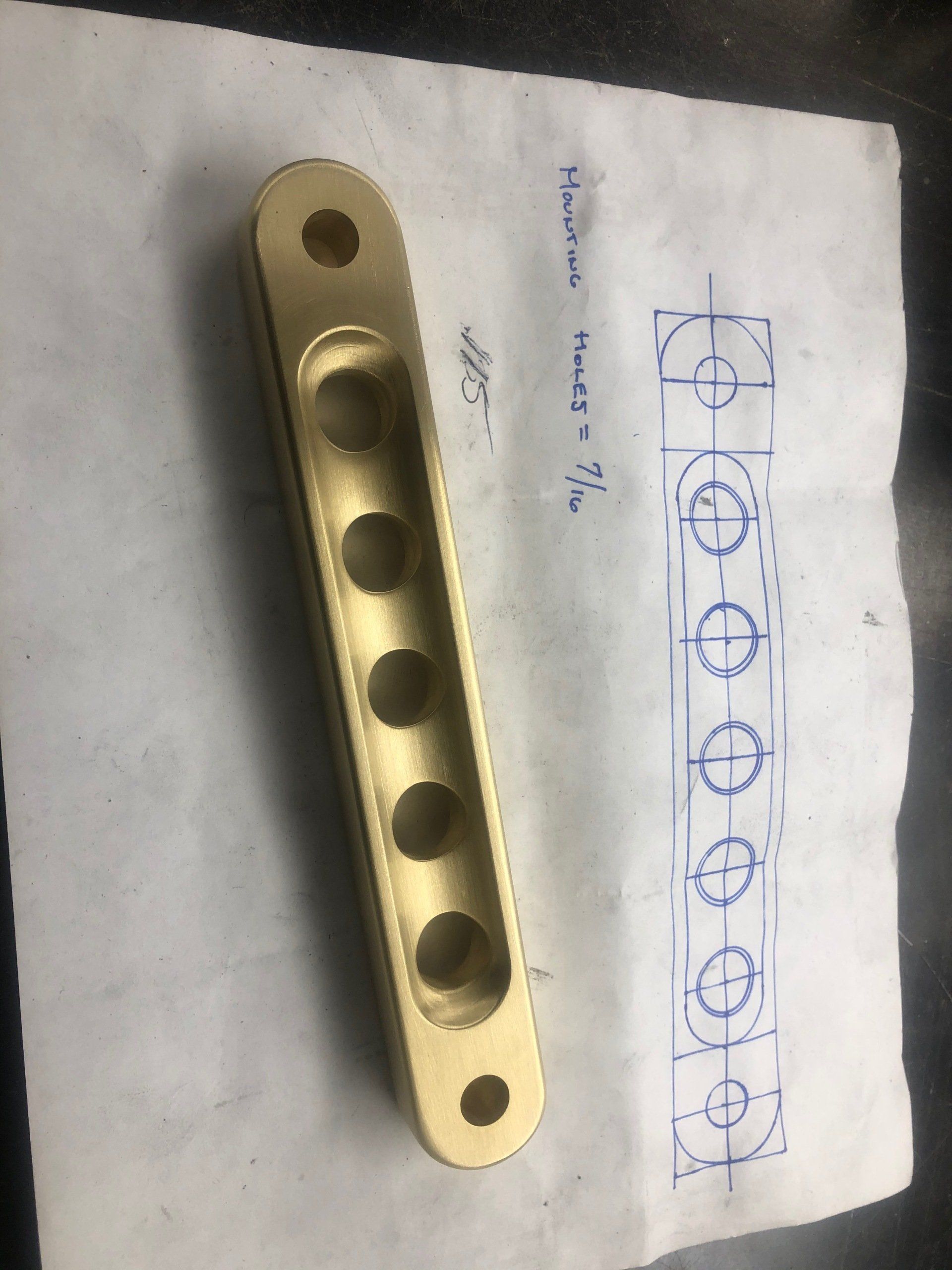 Gold-colored metal mounting bracket with a row of circular holes, shown beside a sketch on paper.