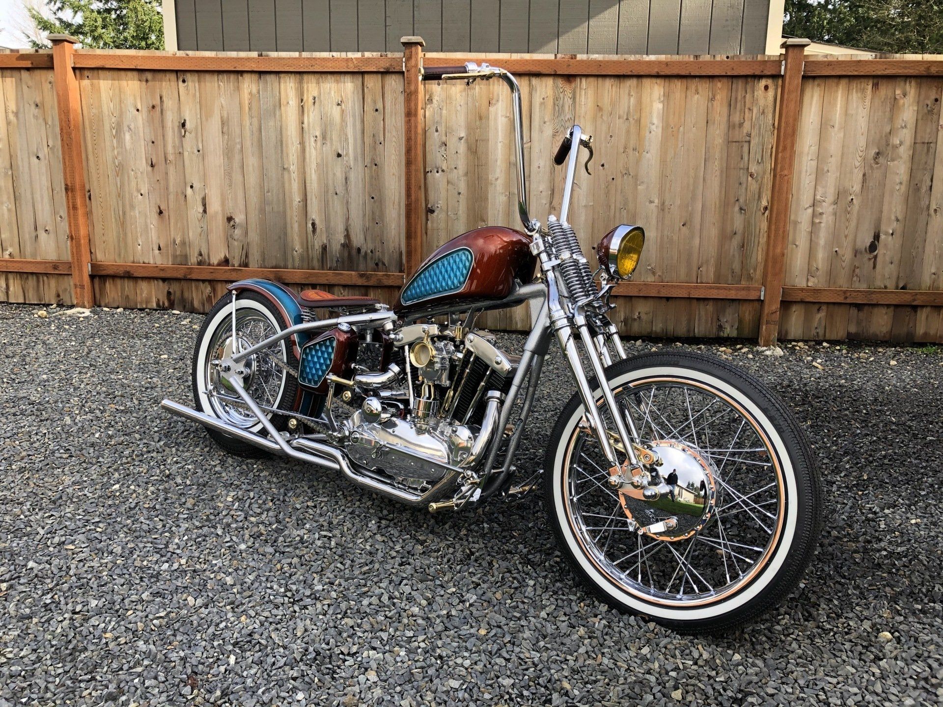 Custom chopper motorcycle parked on gravel in front of a wooden fence