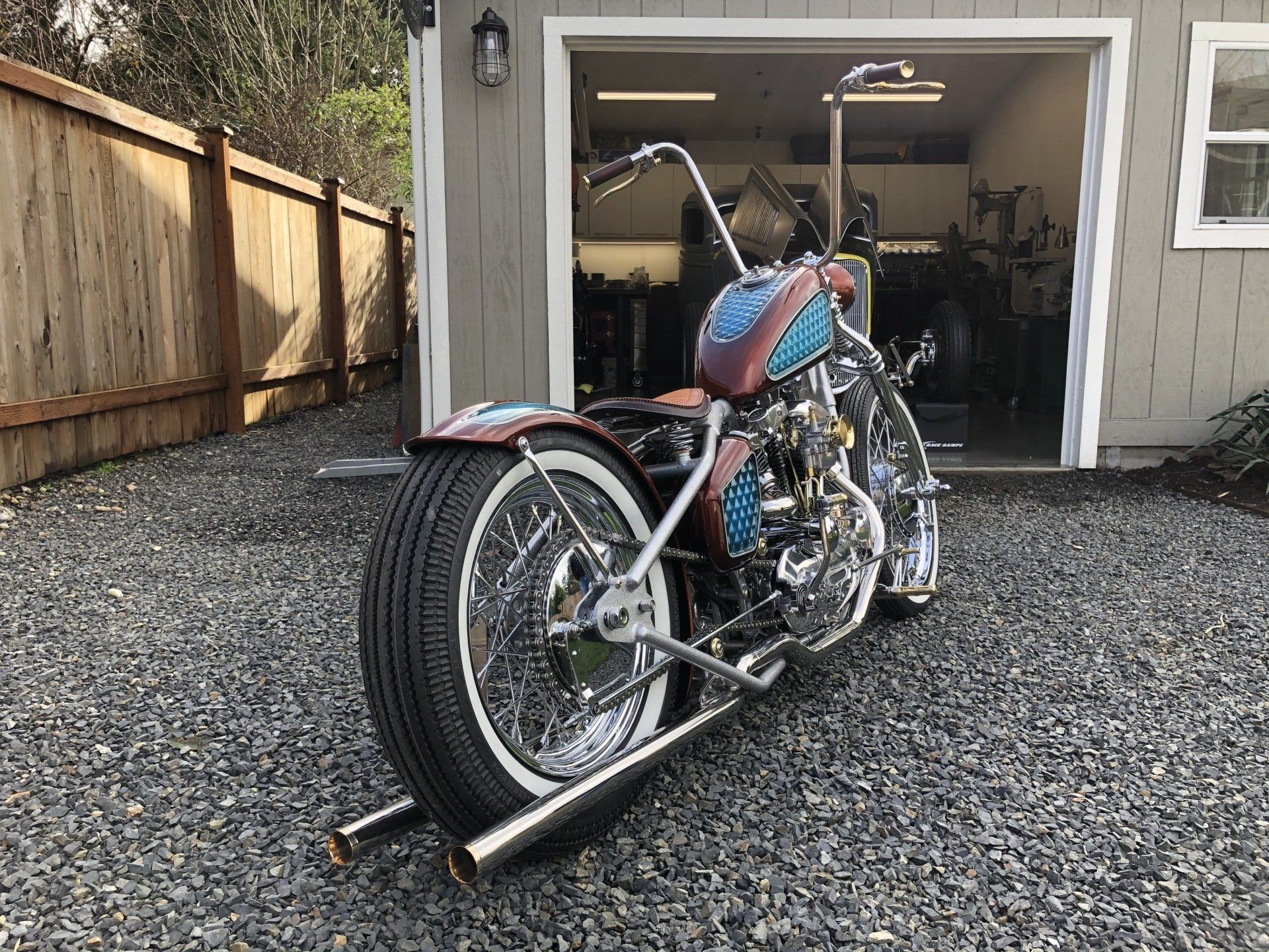 Custom chopper motorcycle parked on gravel in front of an open garage, blue and red tank visible