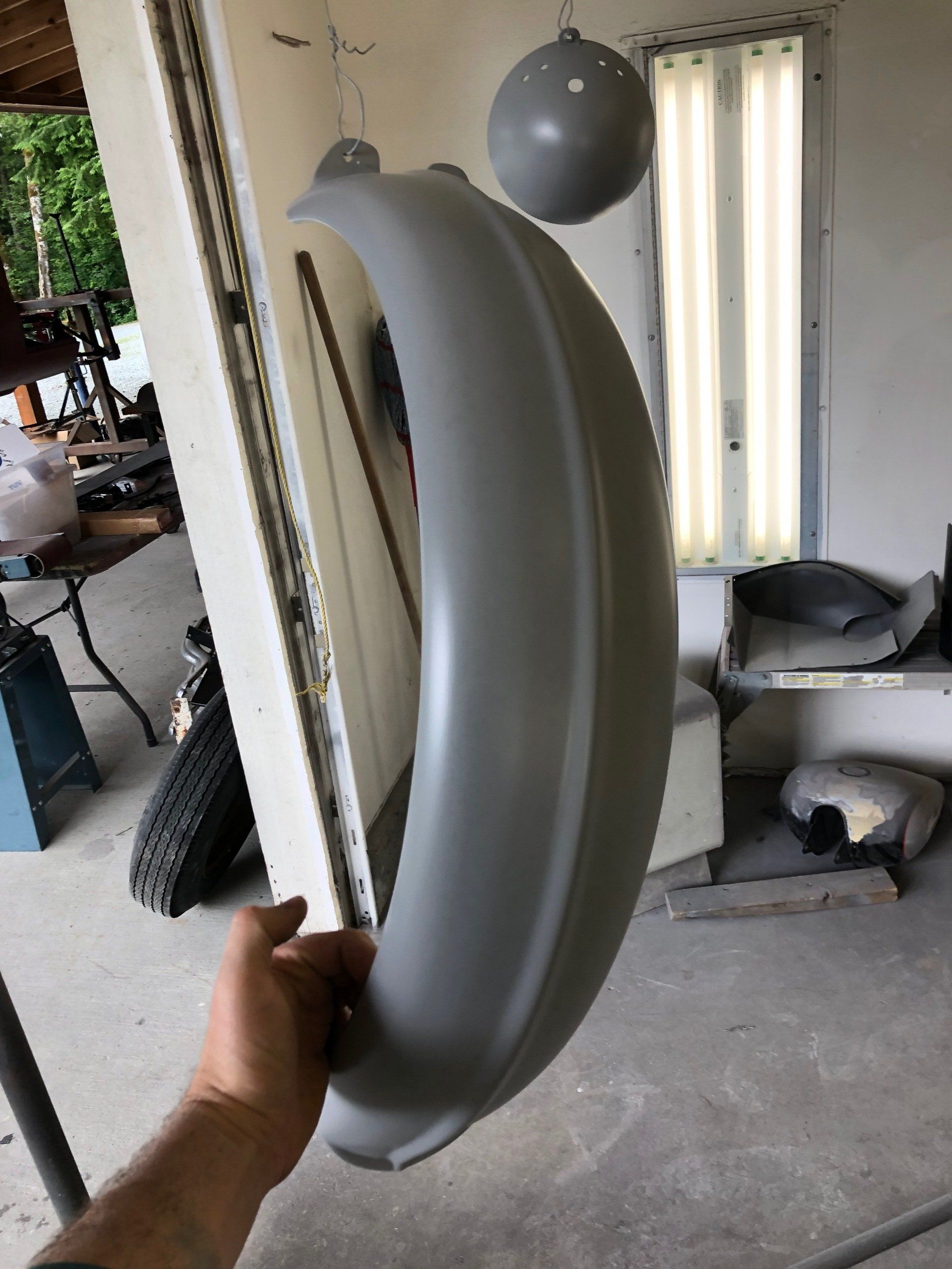 Hand holding a gray plastic car part indoors, with hanging fixtures in the background