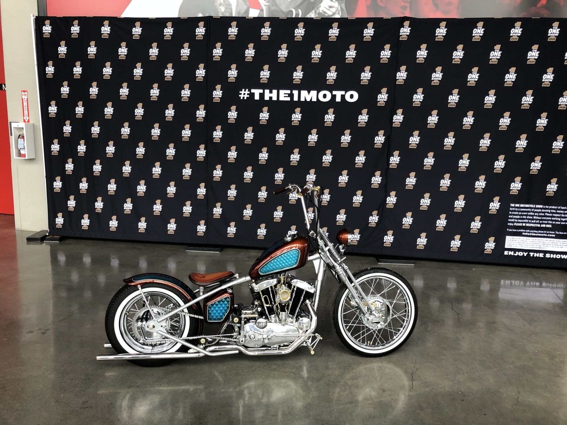 Custom chopper motorcycle with blue tank displayed indoors in front of a THEMOTO backdrop
