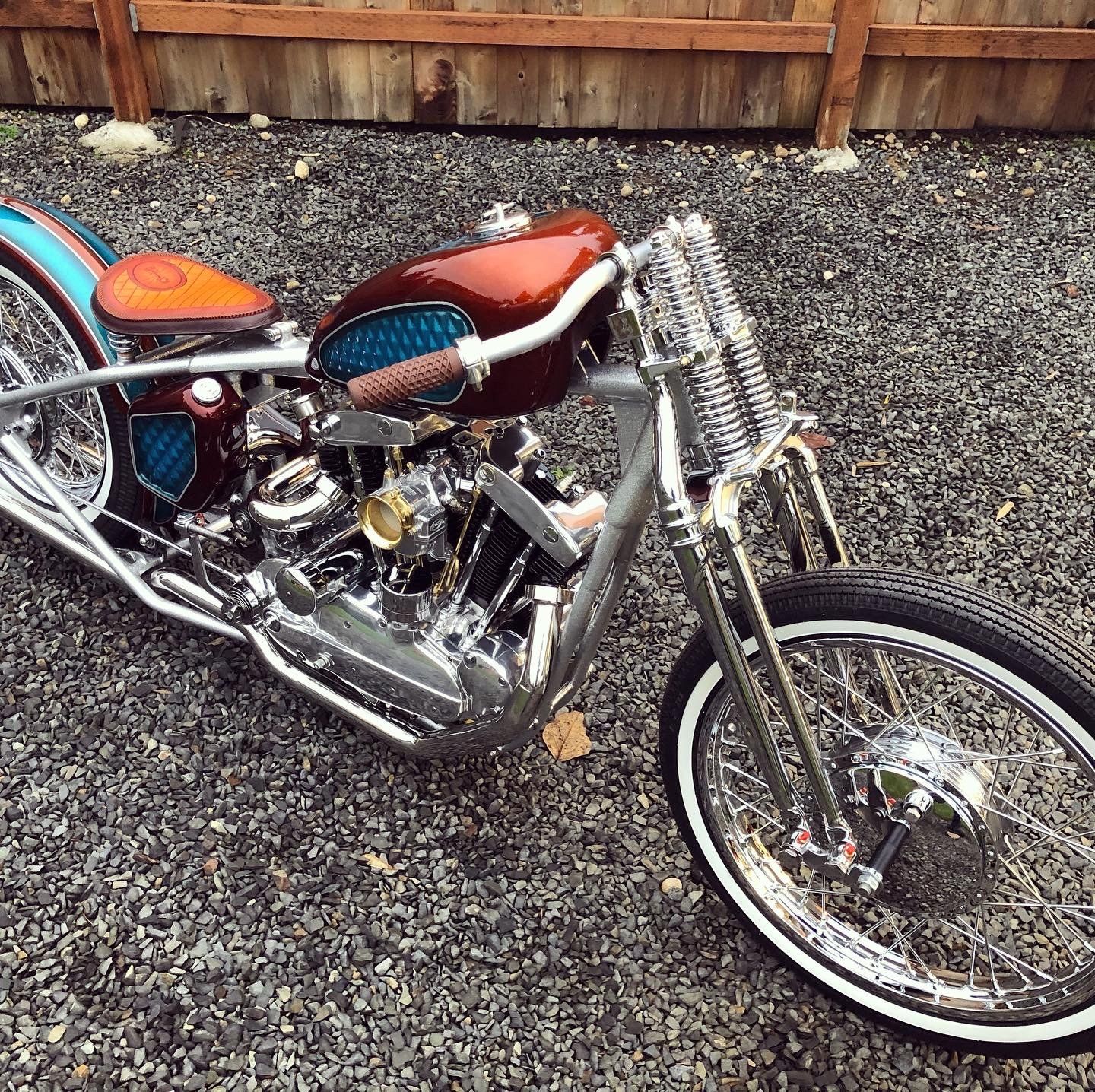 Custom orange-and-chrome chopper motorcycle parked on gravel beside a wooden fence.