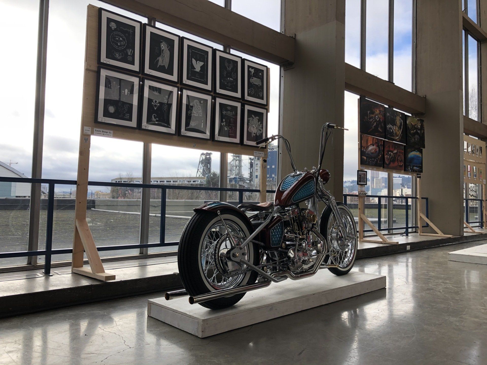 Custom motorcycle on display in a bright gallery with framed photos and large windows overlooking water