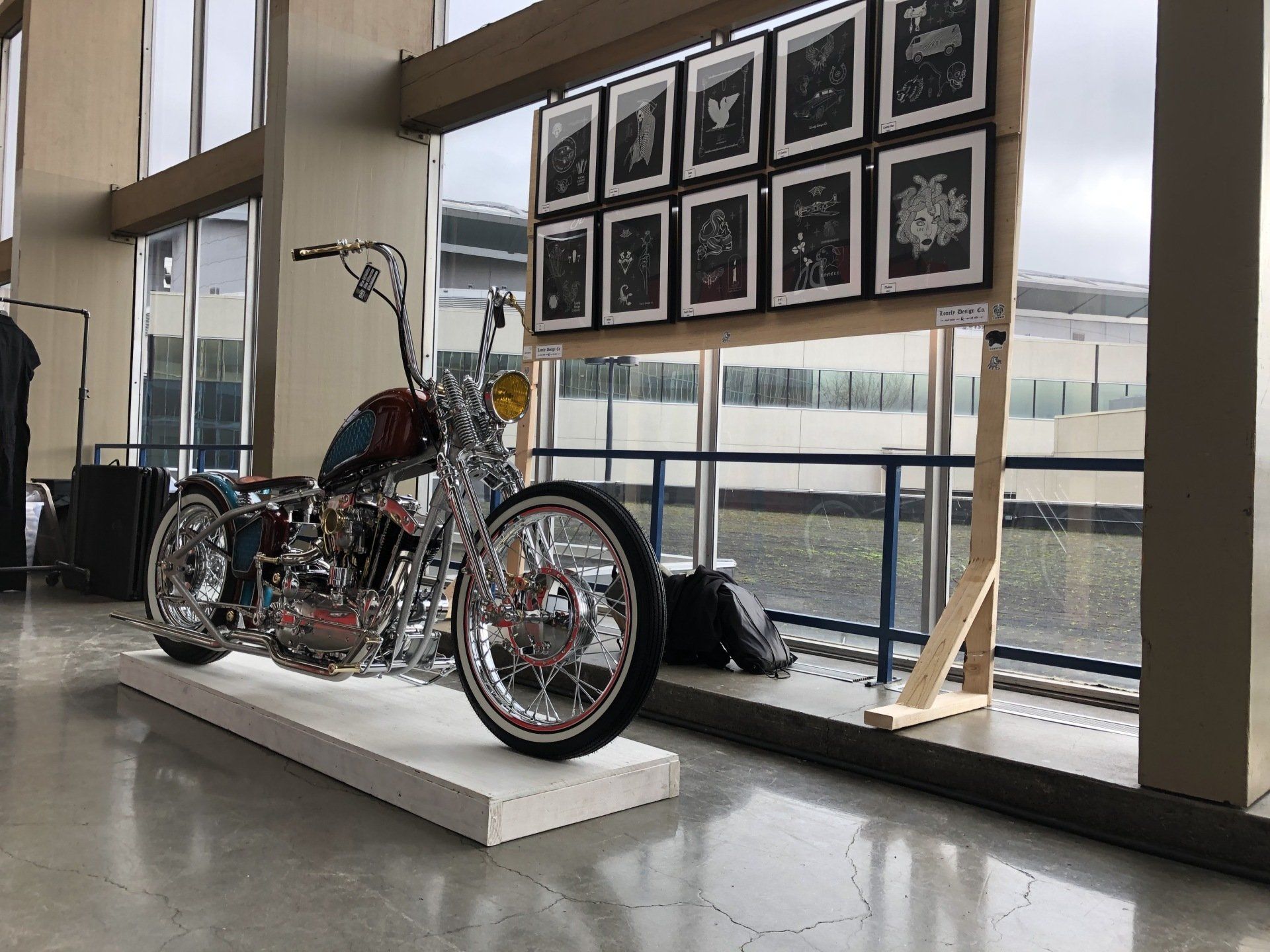Custom three-wheeled motorcycle displayed indoors beside framed photos and large windows.