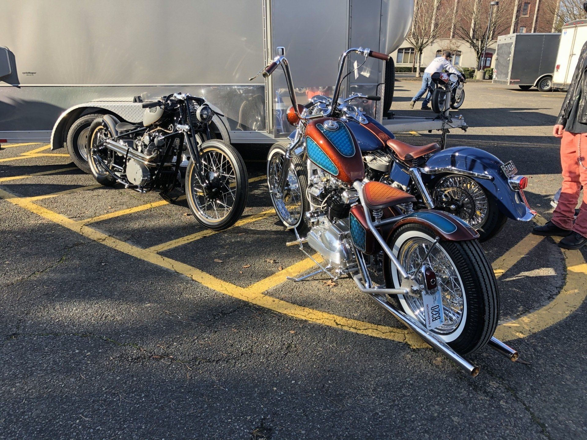 Three custom motorcycles parked in a lot beside a large silver trailer, one blue and orange, two black.