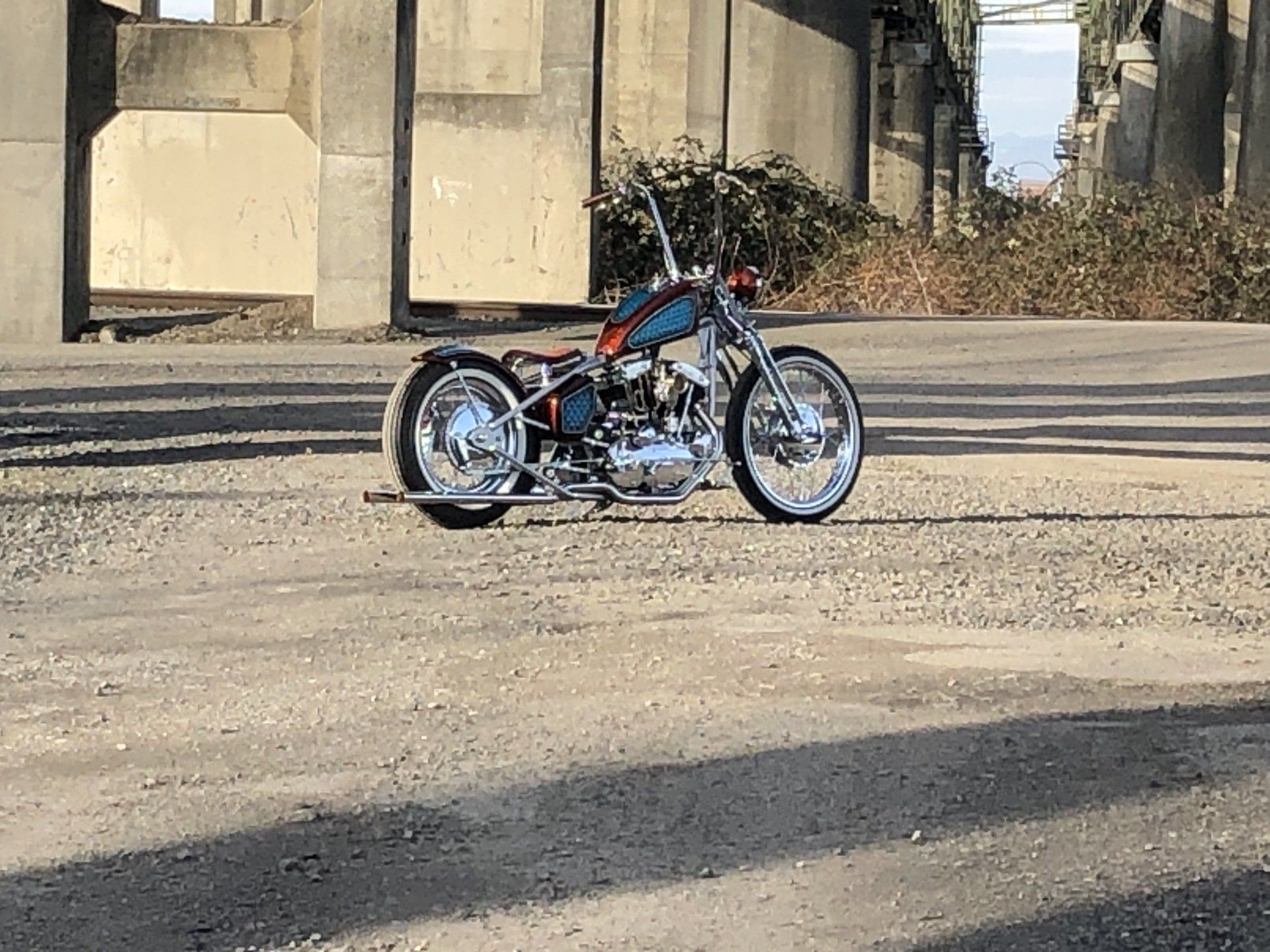 Custom low-slung trike bike parked on a concrete lot beside a bridge column
