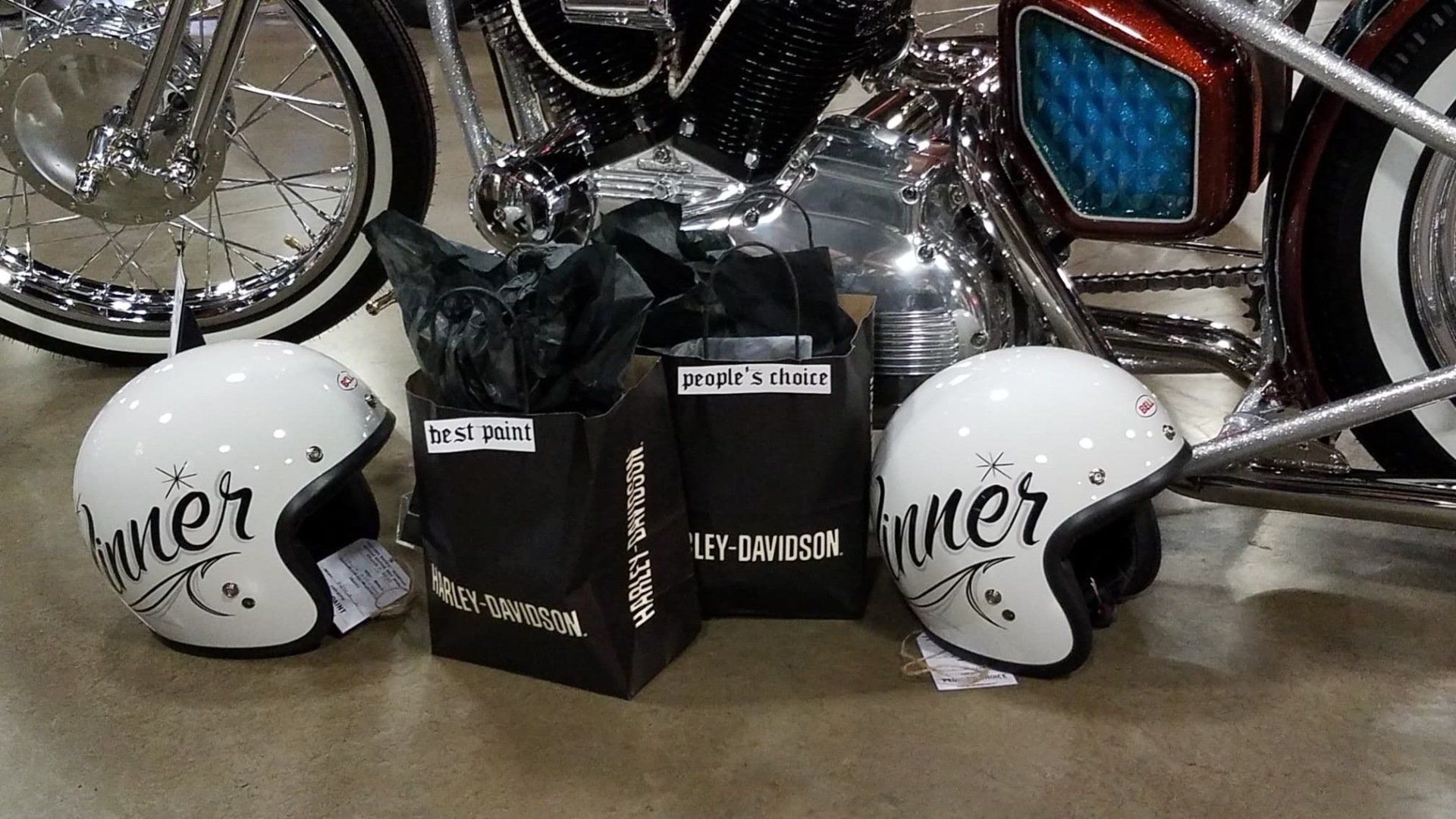 Two white helmets and black gift bags beside a chrome motorcycle, with a bicycle wheel in the background