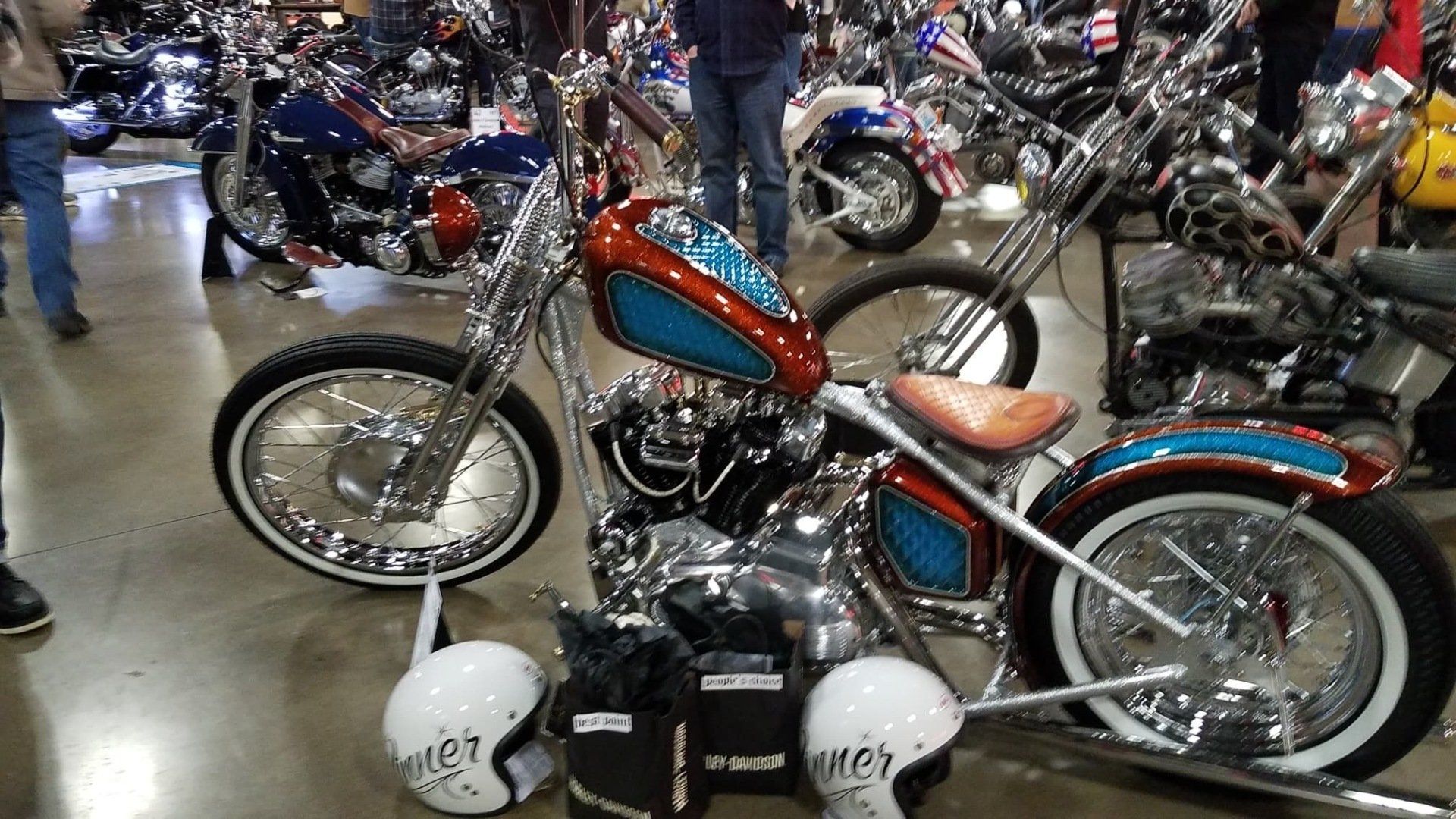Customized orange-and-blue motorcycle displayed indoors among other motorcycles, with white helmets on the floor.