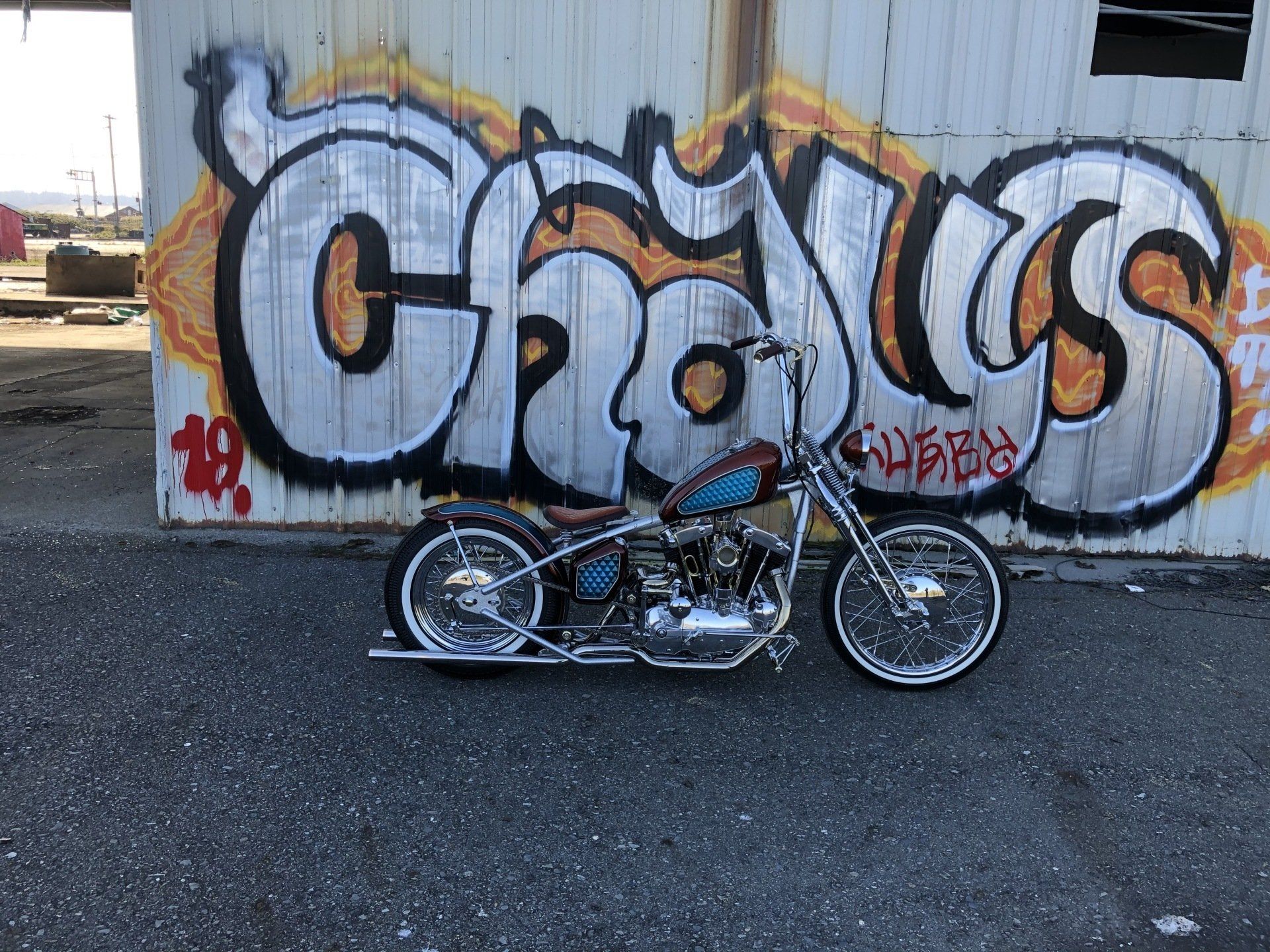 Custom lowrider motorcycle parked in front of orange-and-black graffiti on a wall.