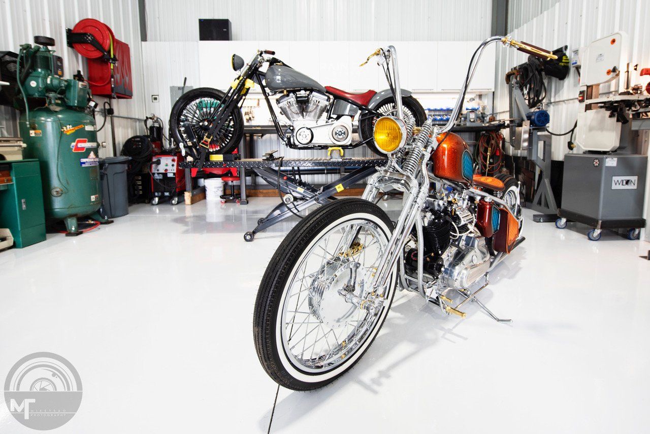 Custom motorcycle in a bright workshop, with exposed chrome frame and a large front wheel in the foreground.