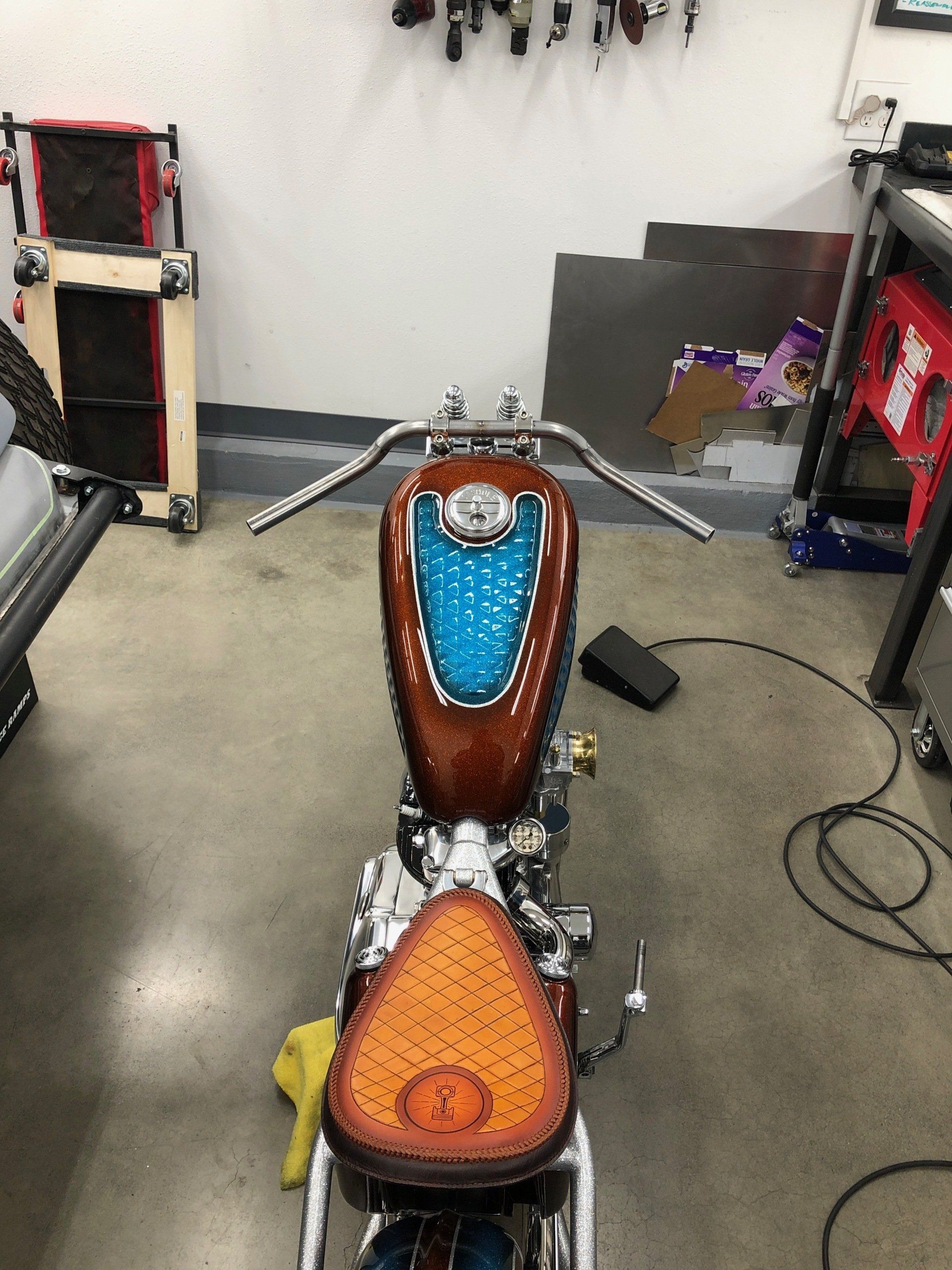 Custom motorcycle viewed from the front in a garage, with blue and orange seats and chrome handlebars.