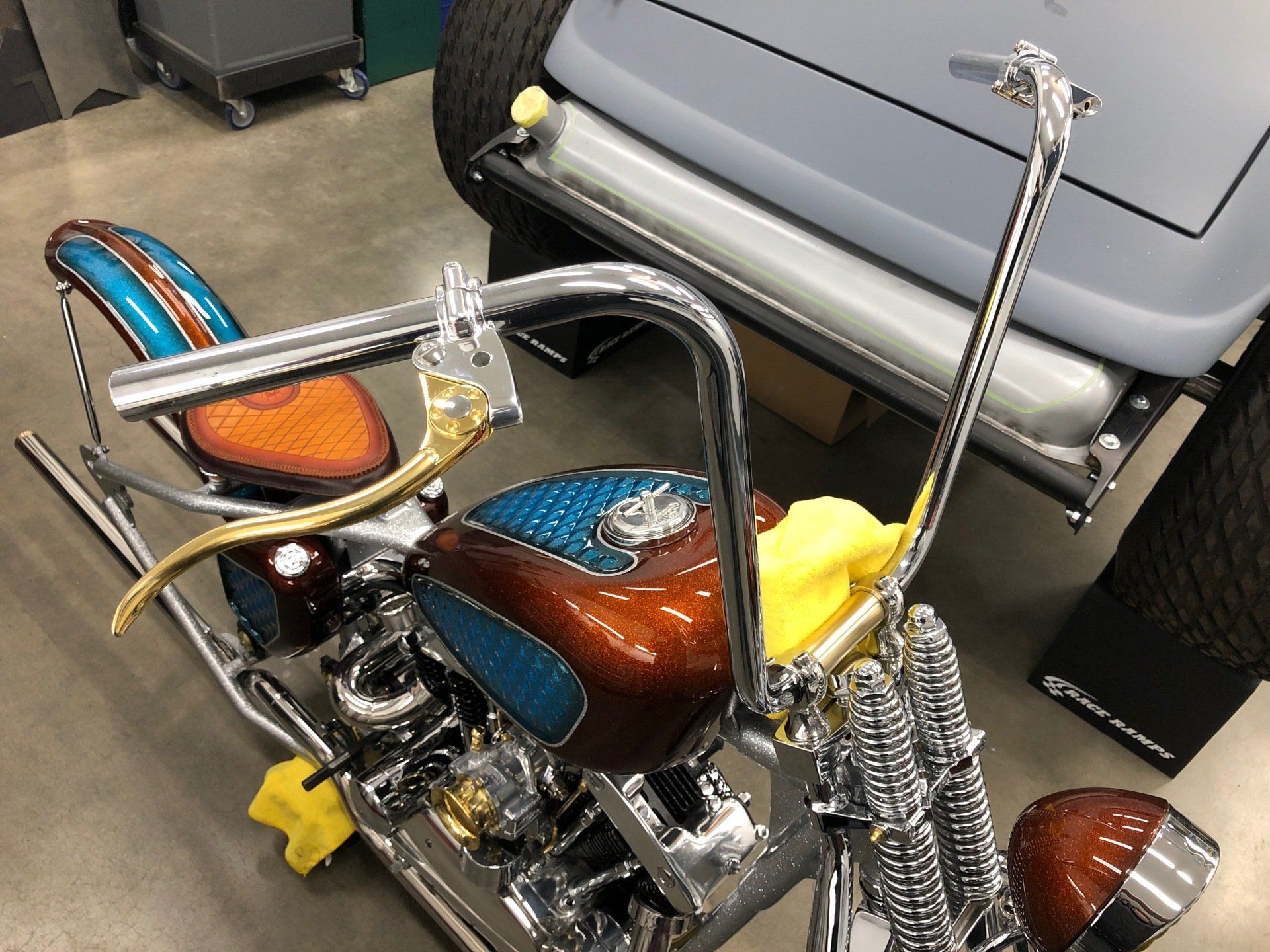 Custom chopper motorcycle with chrome handlebars and brown-blue seat, parked indoors beside a gray vehicle