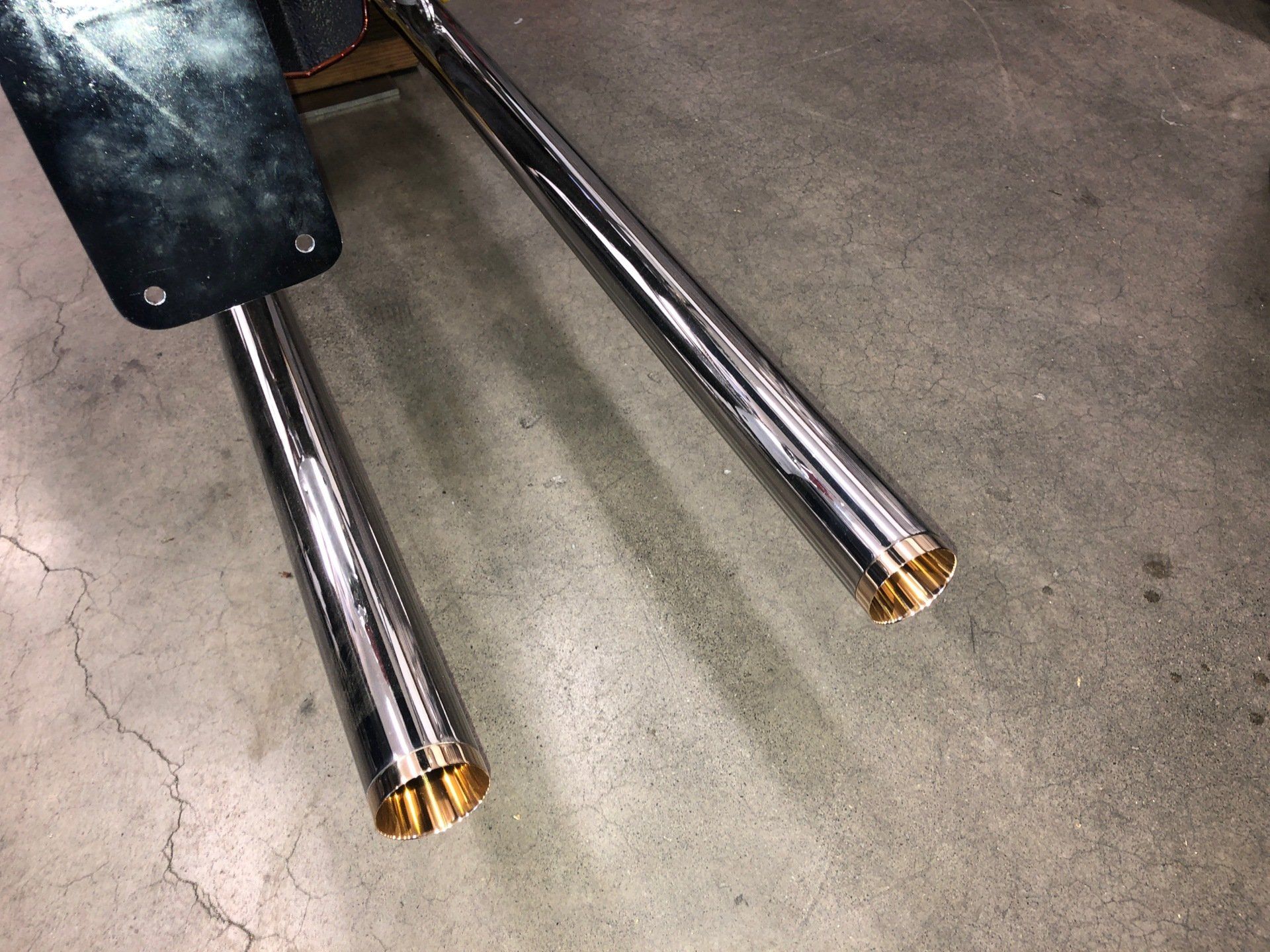 Two chrome exhaust pipes with gold tips extending from a black vehicle body onto concrete.