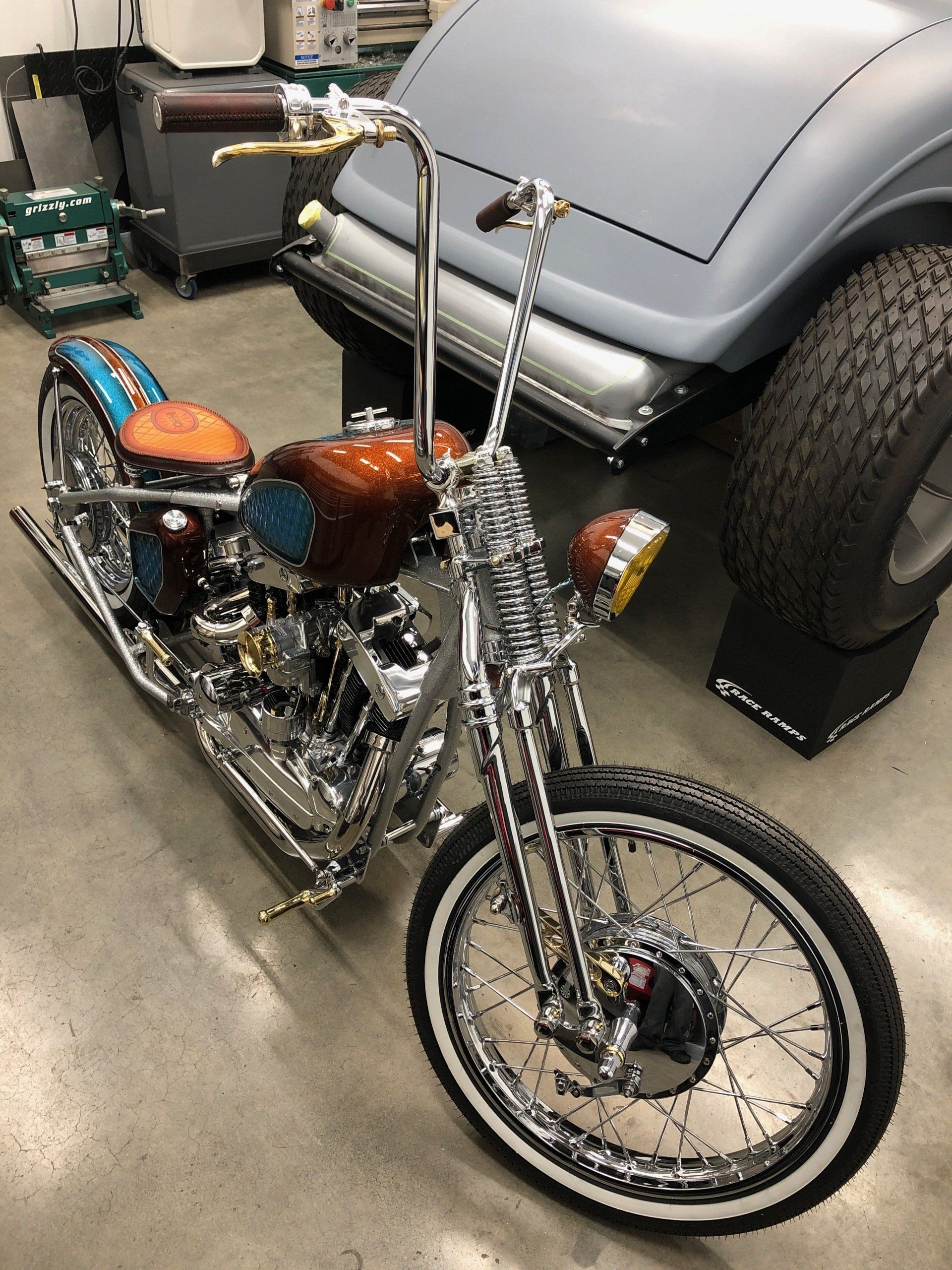 Custom chopper motorcycle with chrome details and orange seat parked in a garage beside a classic car.