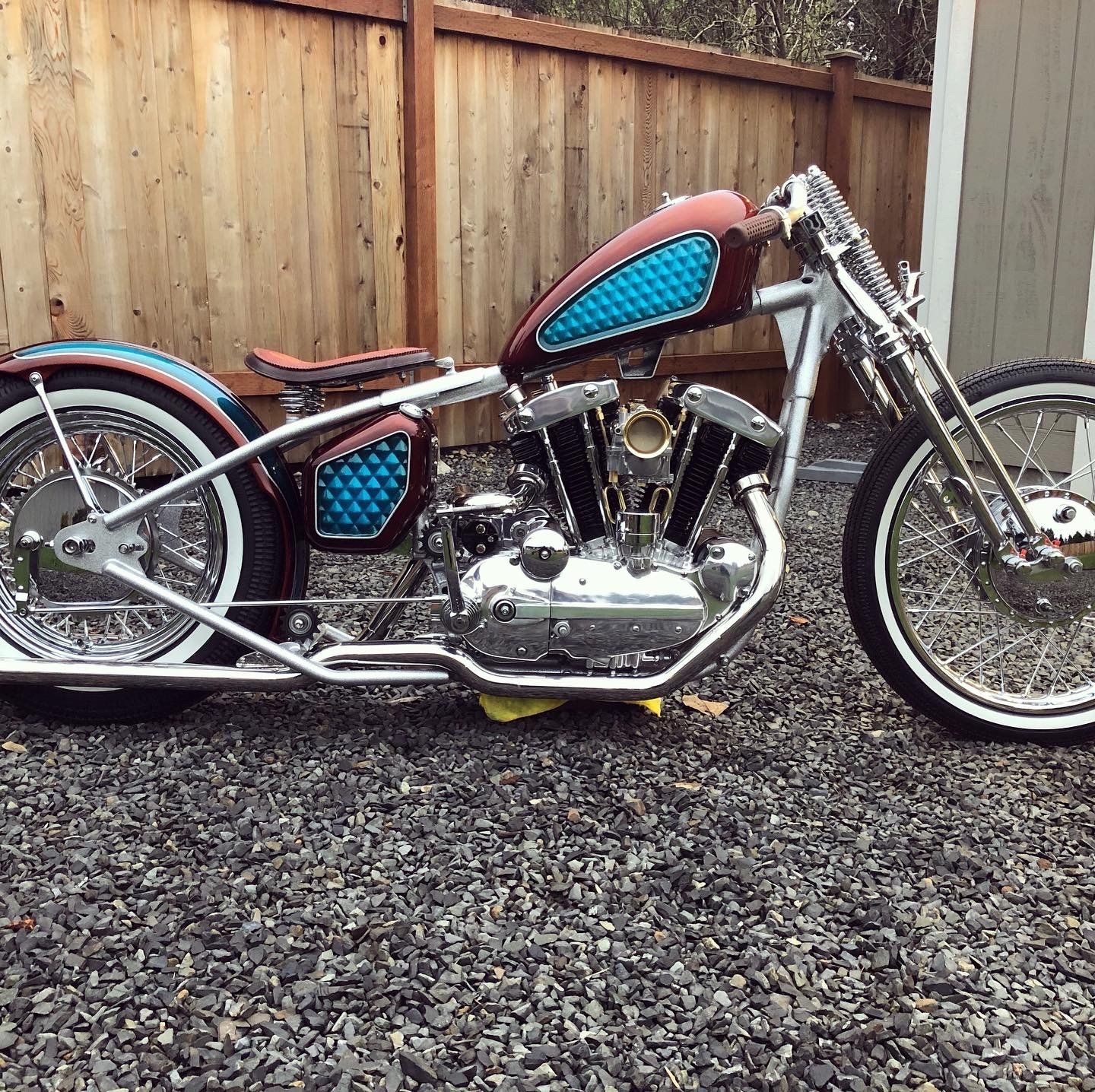 Custom chopper motorcycle with blue seat and red tank parked on gravel by a wooden fence