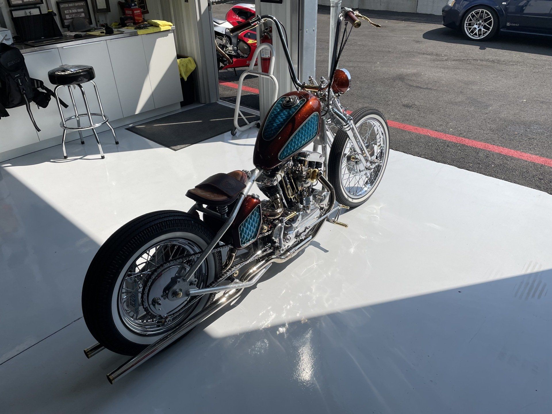 Custom teal-and-orange chopper motorcycle displayed on a white platform at an outdoor event.