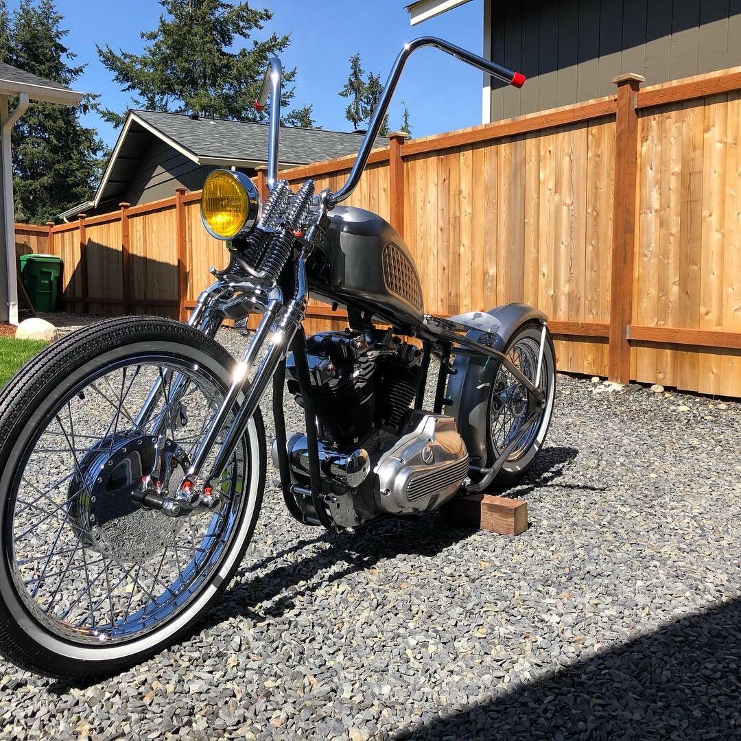 Custom black motorcycle parked on gravel beside a wooden fence in bright sunlight