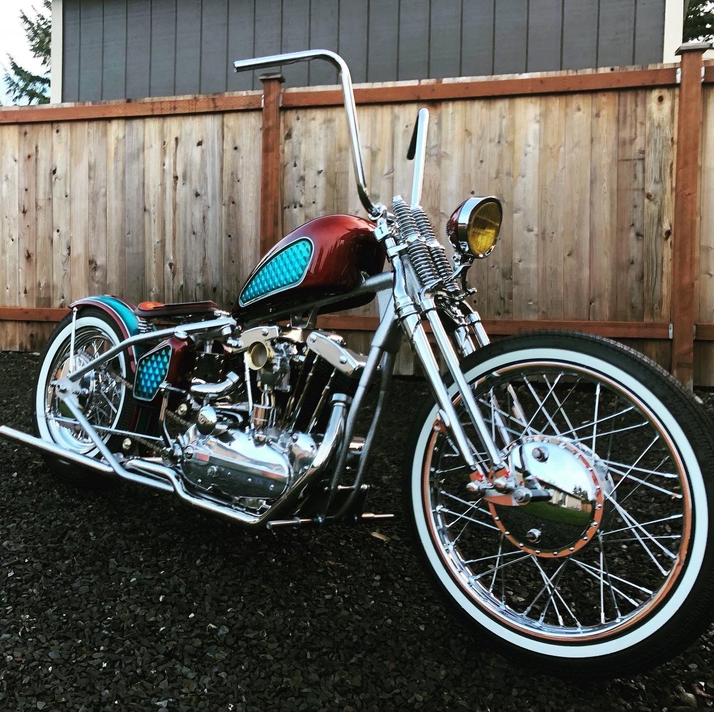 Custom chopper motorcycle with turquoise and red paint parked by a wooden fence outdoors