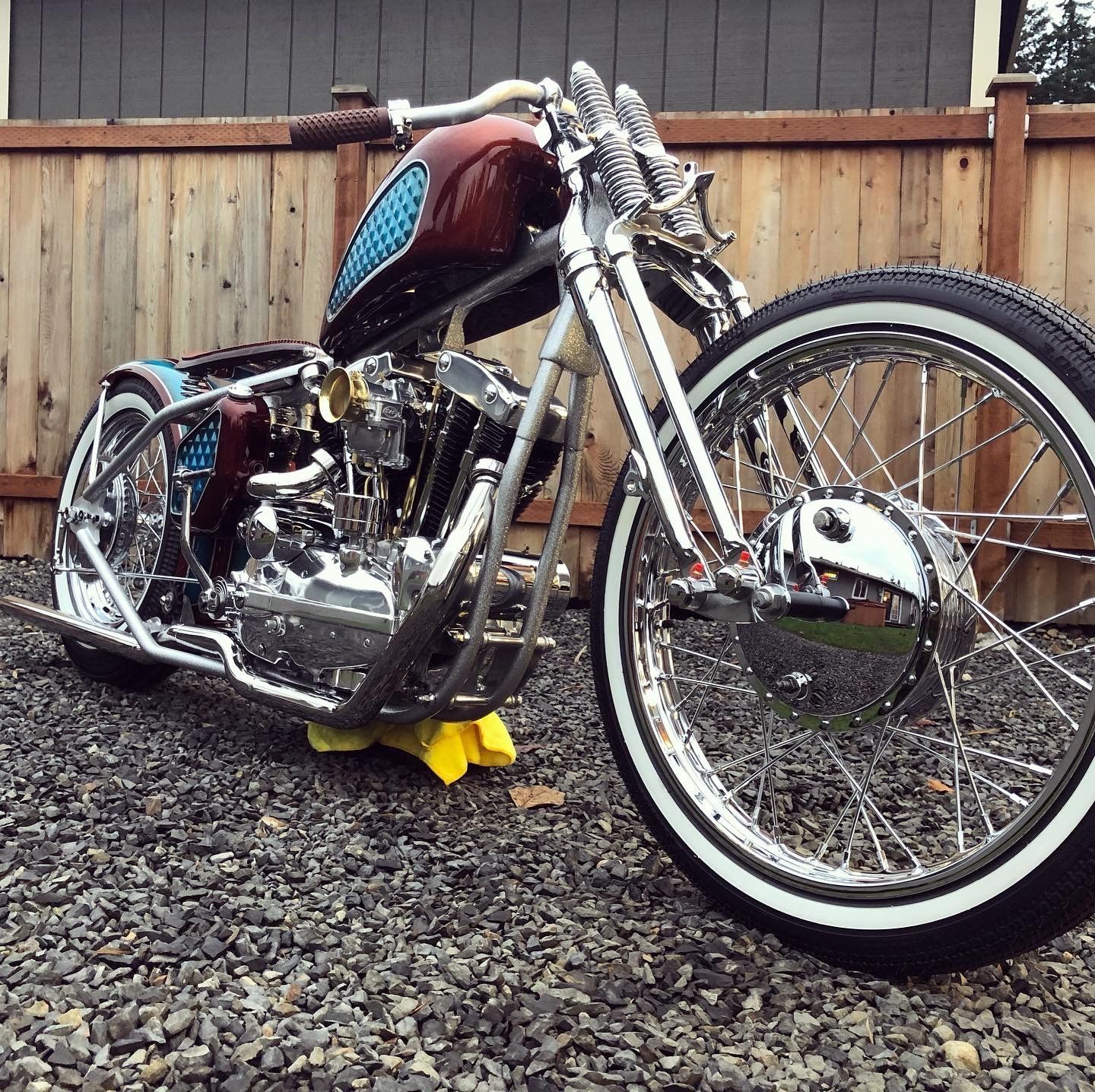 Custom chrome chopper motorcycle with red tank parked on gravel against a wooden fence
