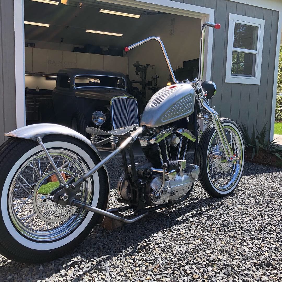 Custom silver chopper motorcycle with whitewall tires parked on gravel in front of a garage