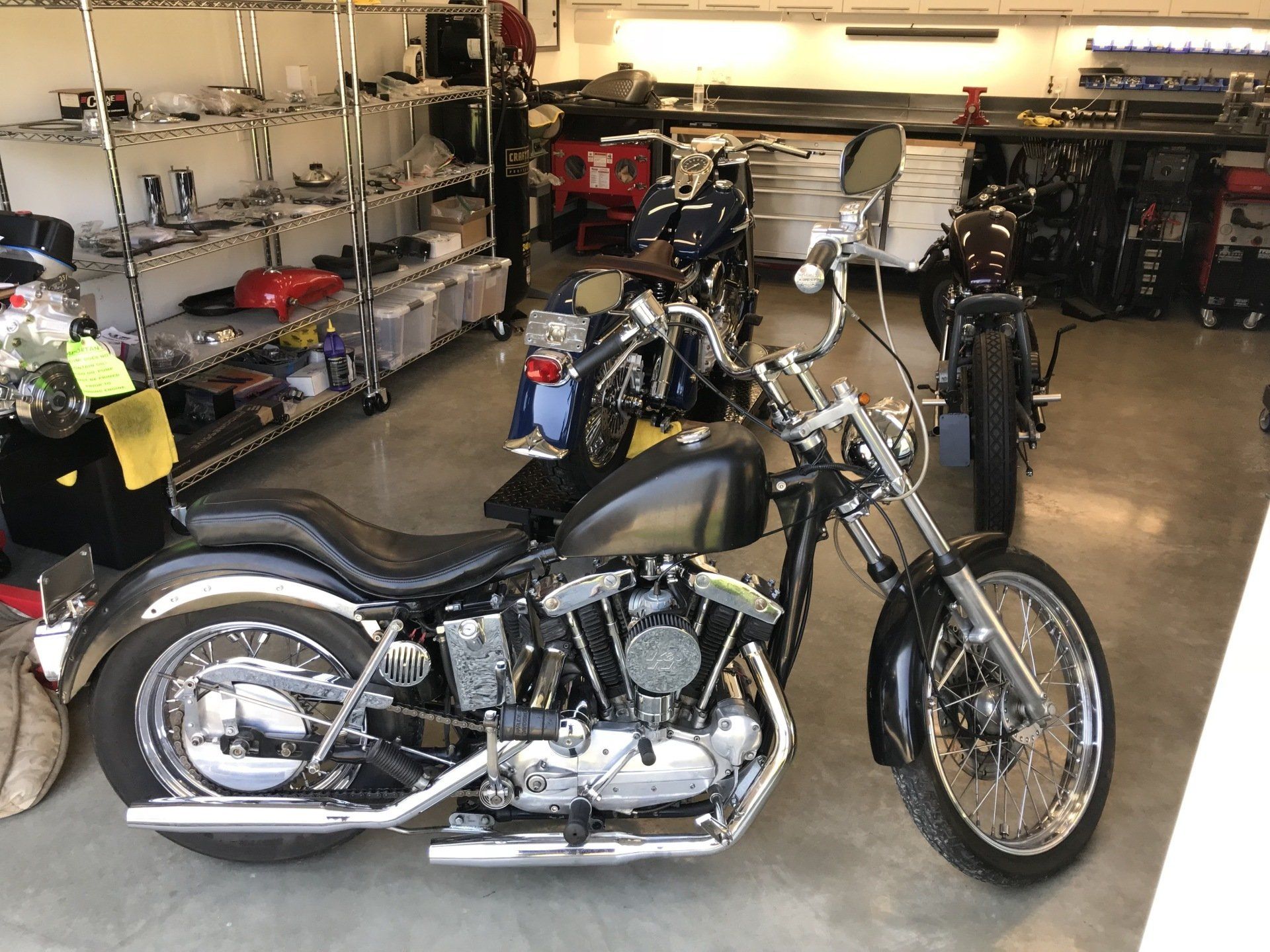 Black custom motorcycle in a garage, with shelves of parts and tools in the background
