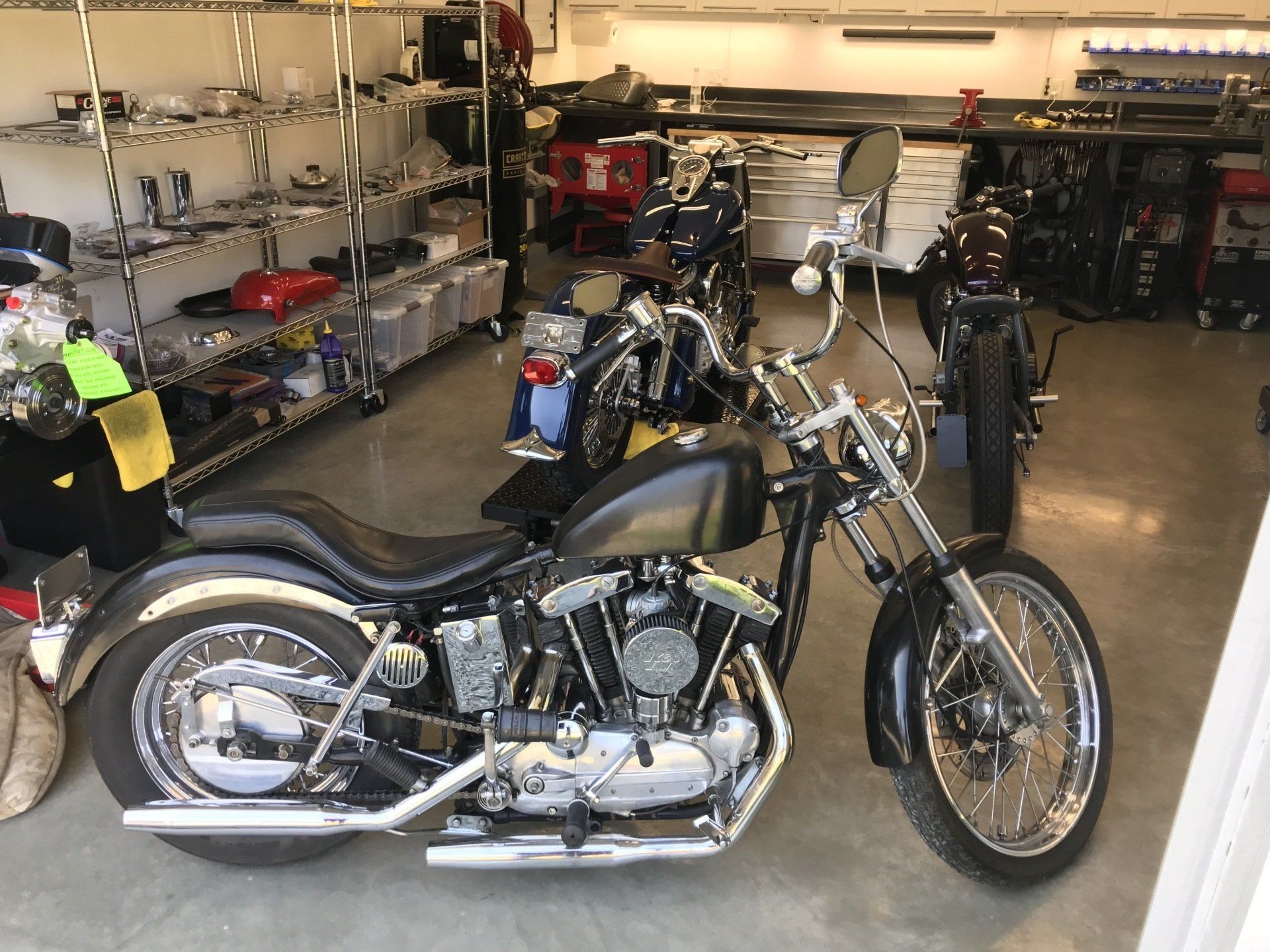 Black motorcycle in a garage or workshop with shelves of tools and parts in the background