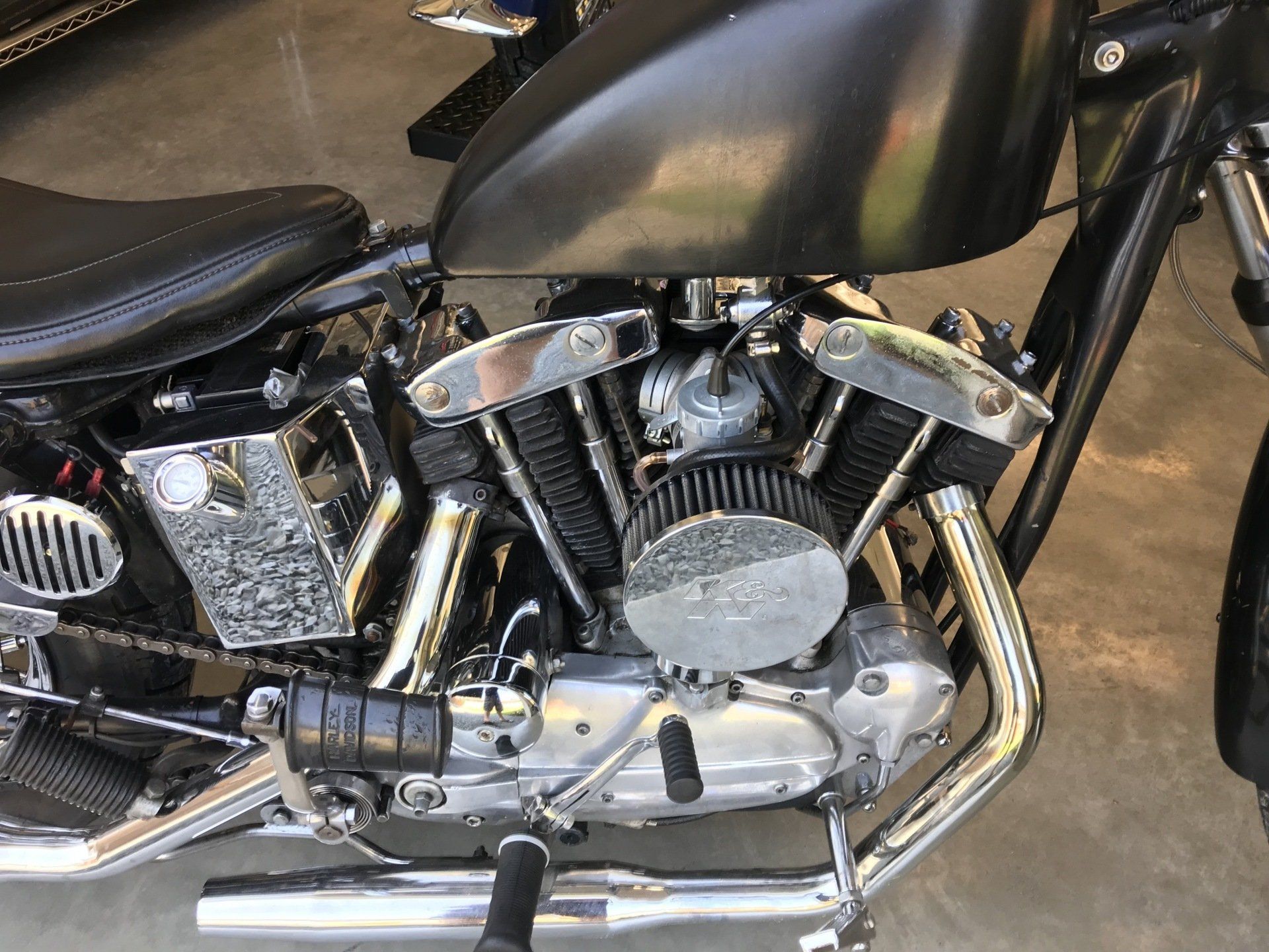Black motorcycle engine and chrome exhaust beneath a black fuel tank indoors