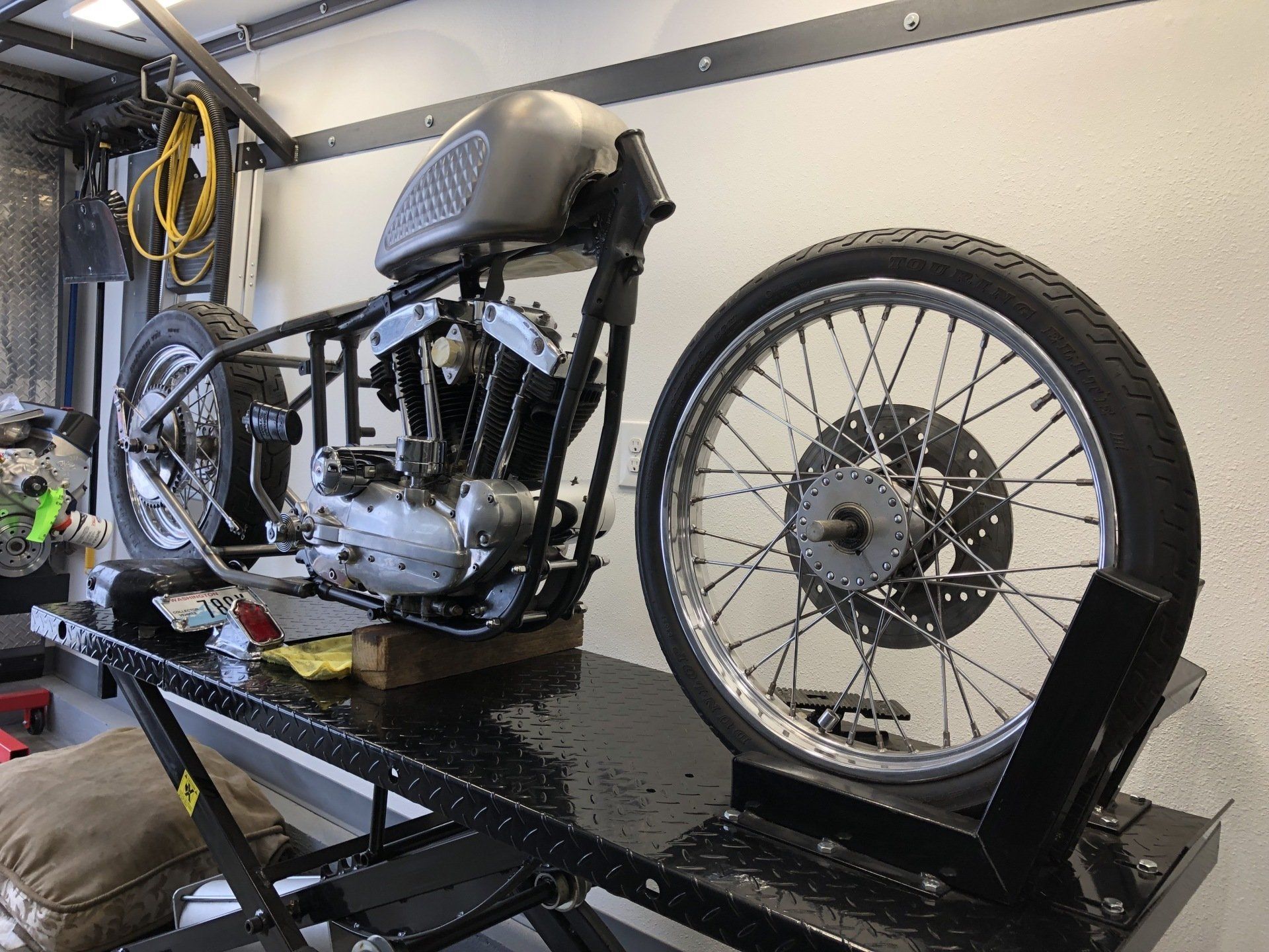 Custom motorcycle frame and engine on a workshop stand, with a rear wheel mounted in the background.