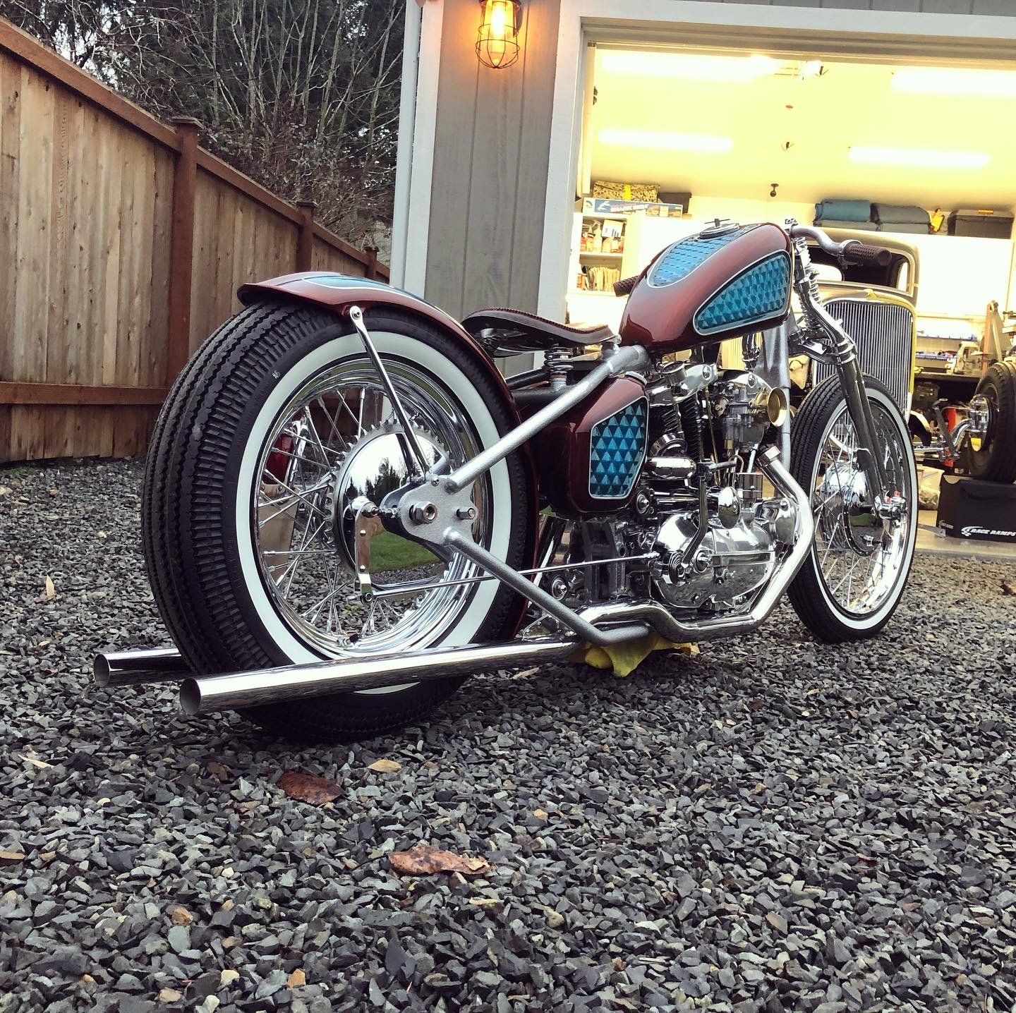 Custom motorcycle with teal tank parked on gravel beside a garage and wooden fence