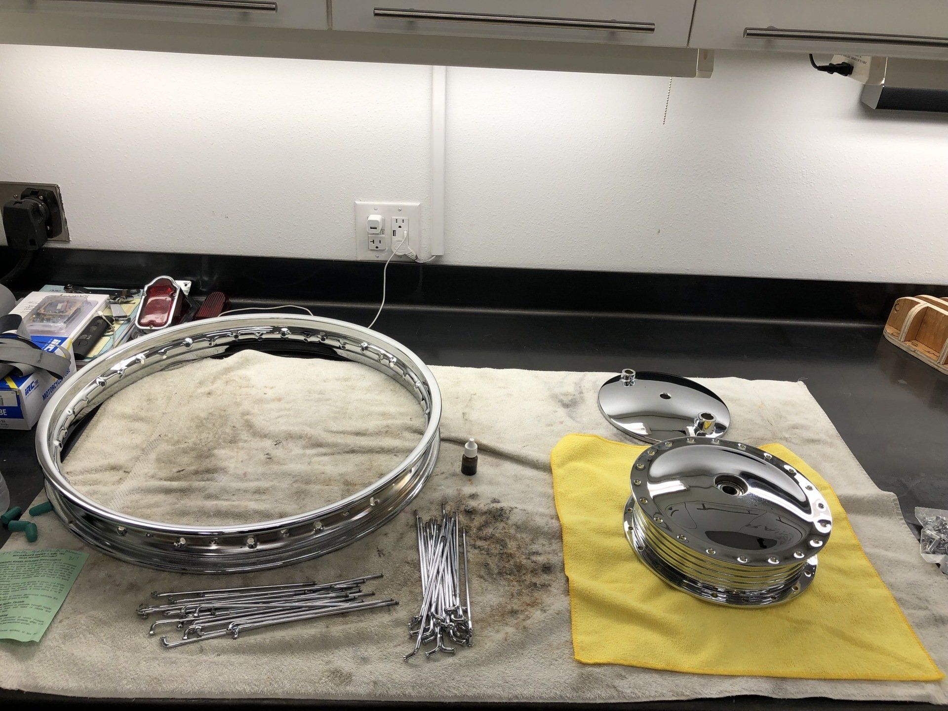Metal drum parts laid out on a workbench, including a large hoop, small hoops, and hardware on a yellow cloth.