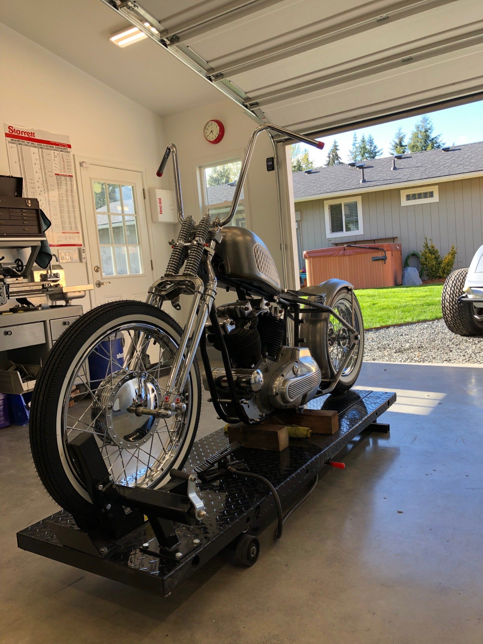 Custom black motorcycle on a lift in a garage, with an open door to a sunny backyard.