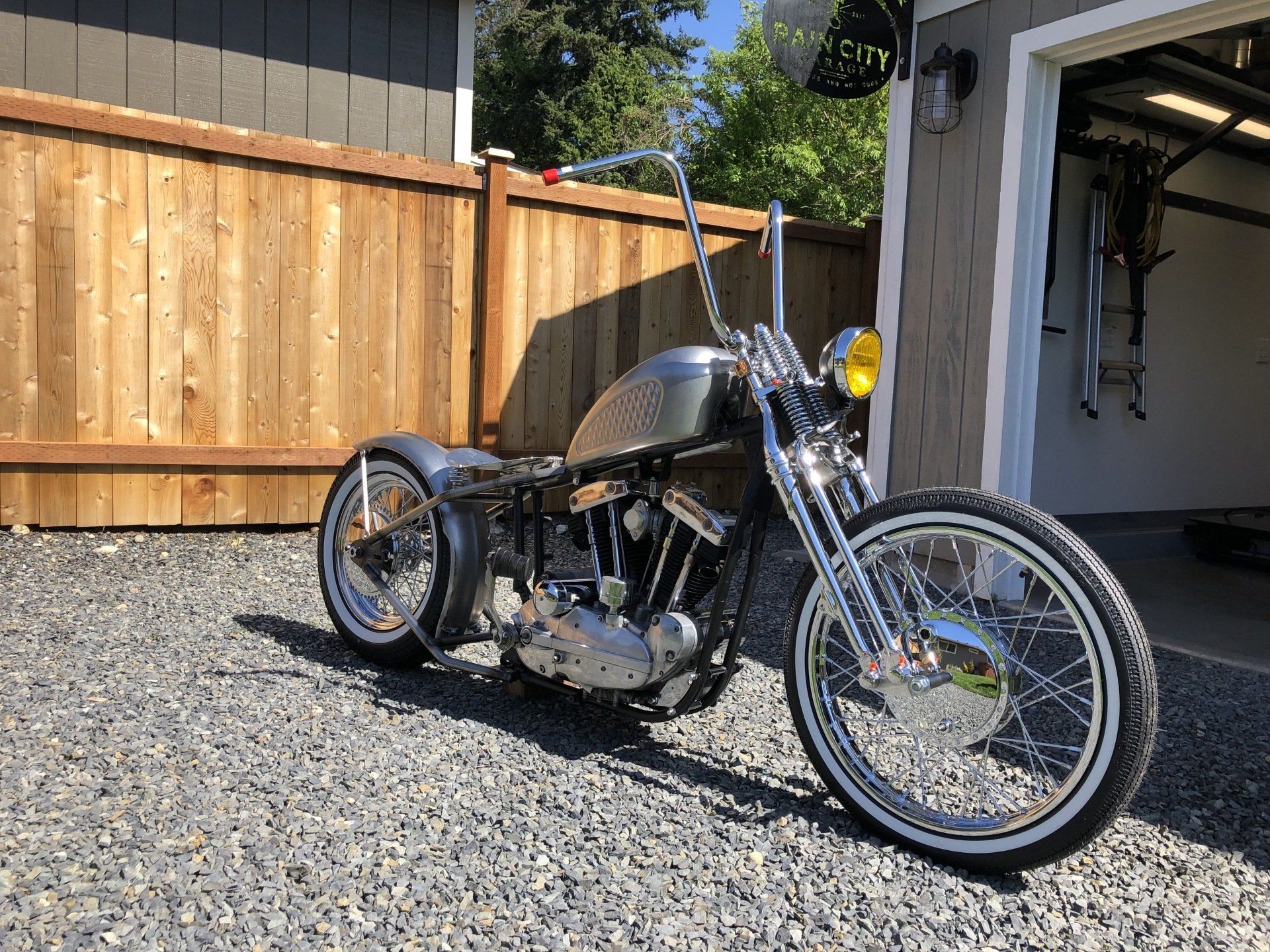 Custom chopper motorcycle parked on gravel in a driveway beside a wooden fence and garage