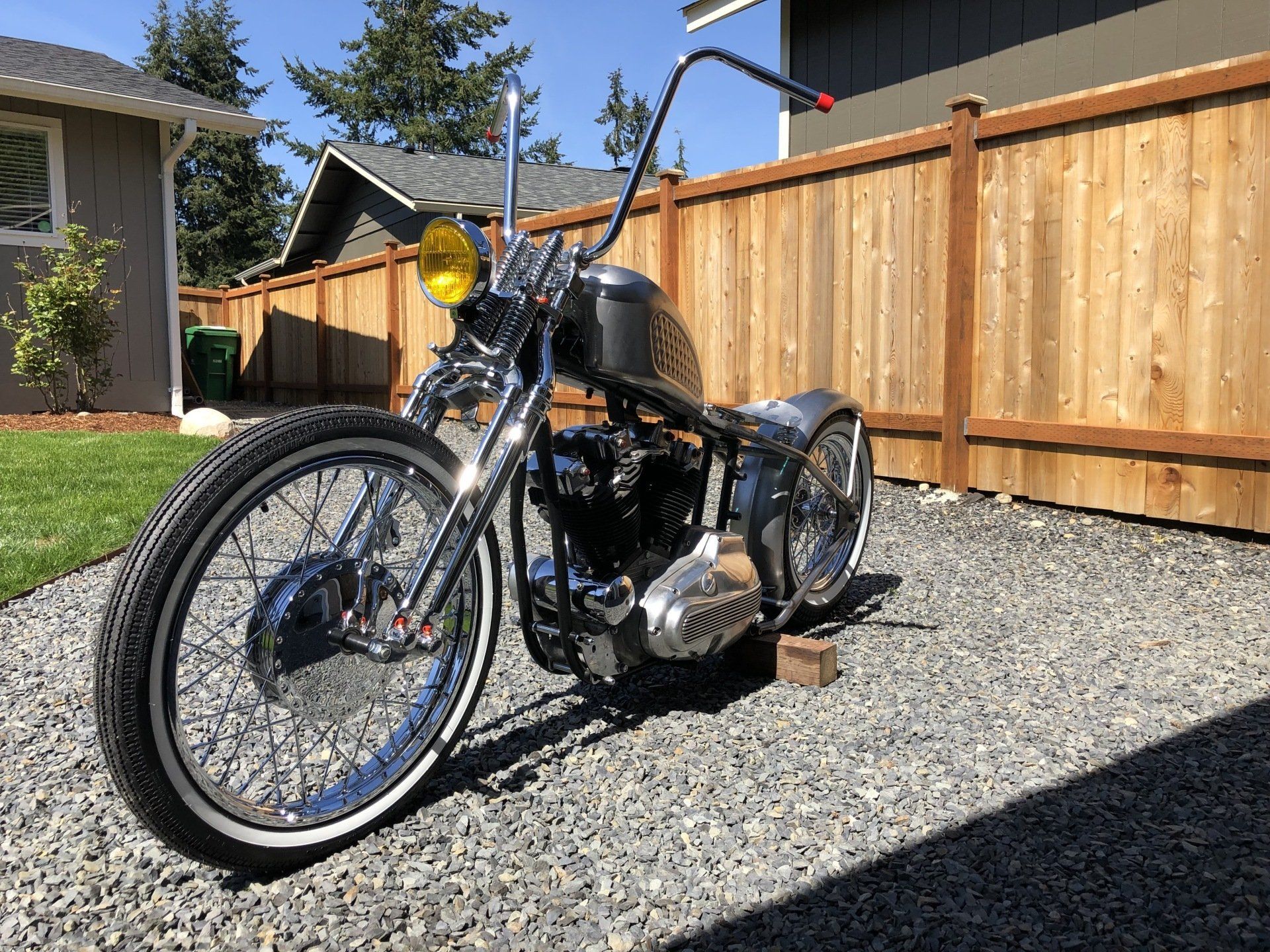 Custom black motorcycle parked on gravel beside a wooden fence in a sunny yard.