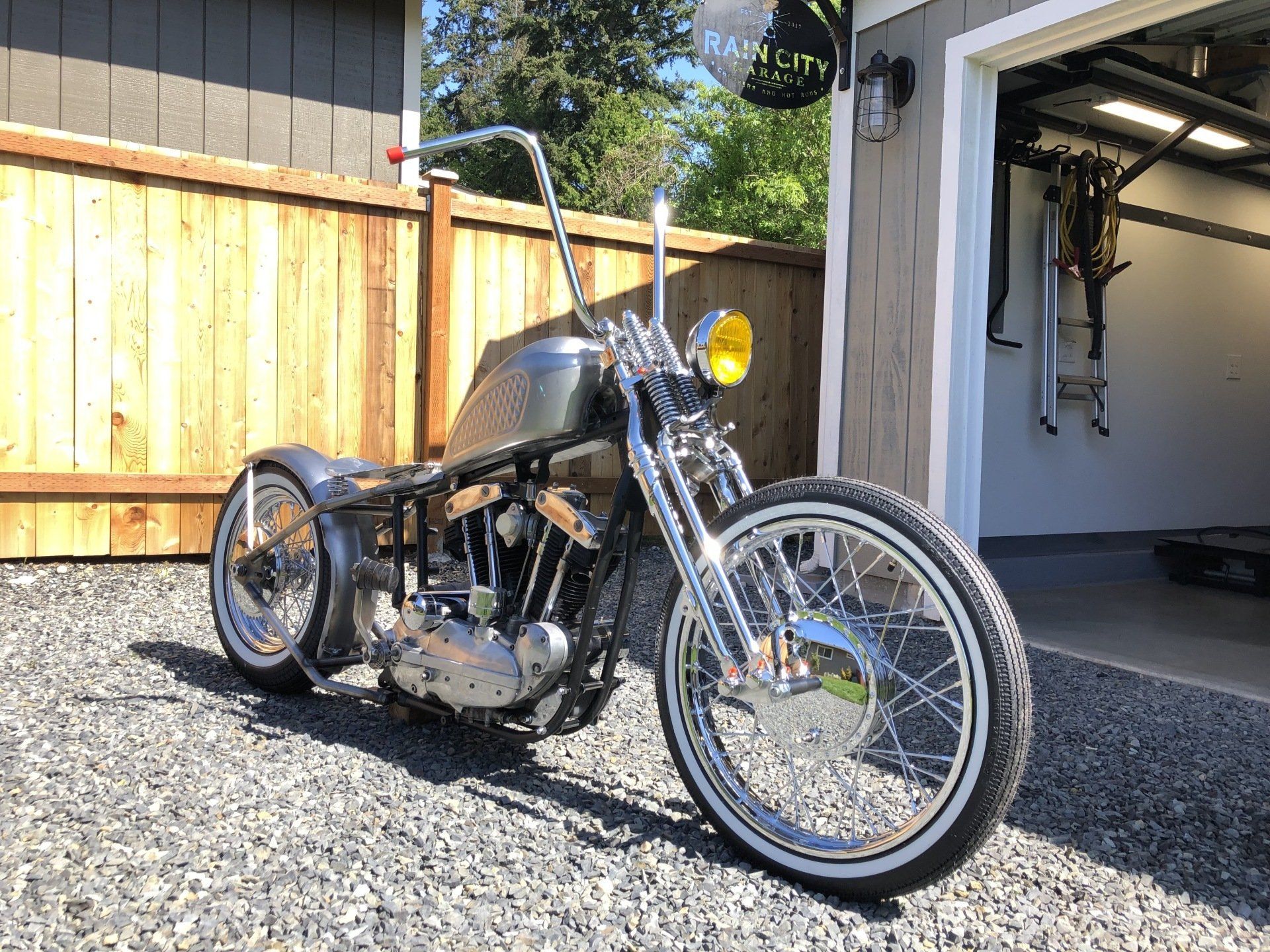 Black custom motorcycle parked on gravel beside a garage, with a wooden fence in the background
