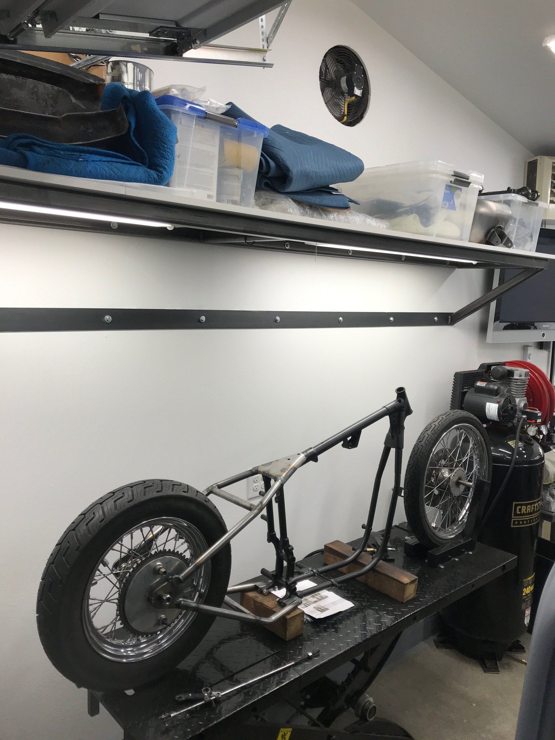 Garage with a custom bicycle frame on a workbench, large wheels attached, and shelves with bags above.
