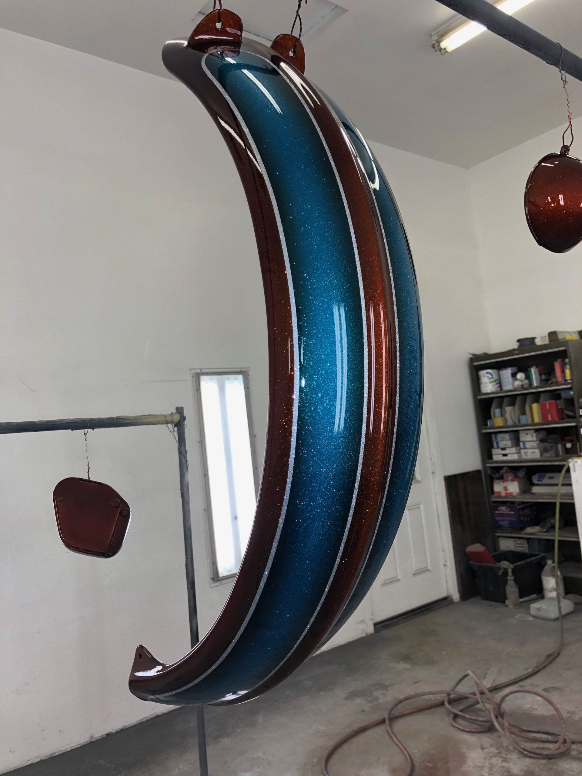 Curved blue-and-brown hanging sculpture in a workshop with shelves and tools in the background