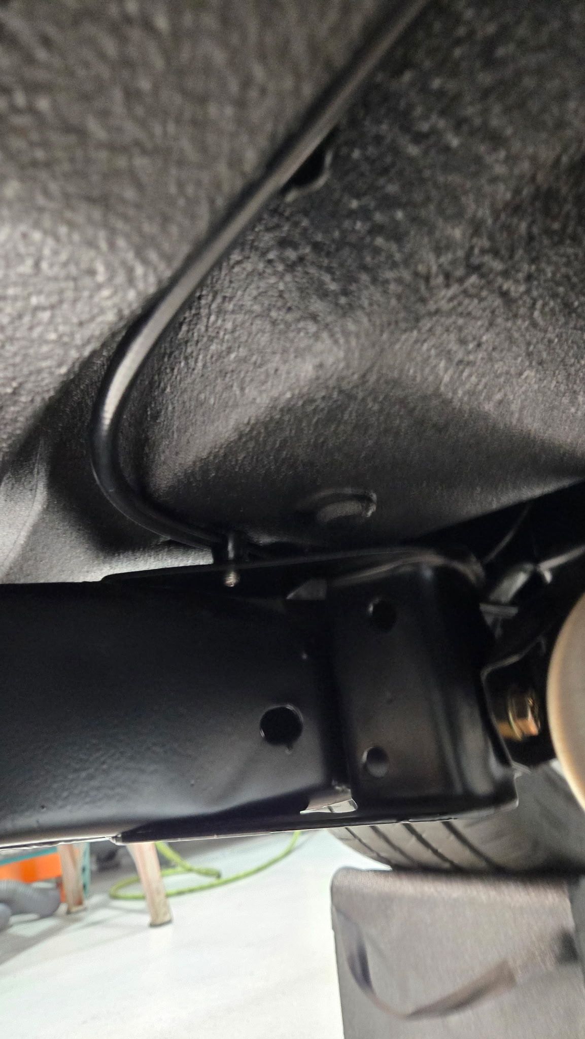 Close-up of a black metal bracket under a vehicle, with textured underbody insulation visible above.