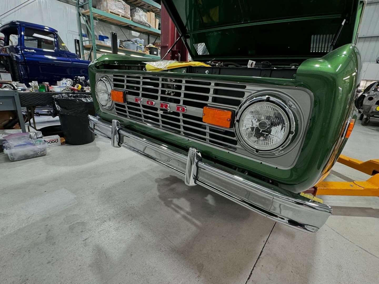 Green classic truck front in a workshop, showing chrome grille and bumper with orange tags.