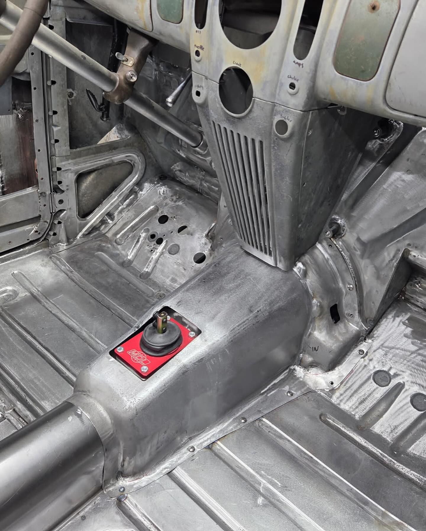 Car interior floor with a red-mounted lever or switch near the center tunnel and exposed metal panels.