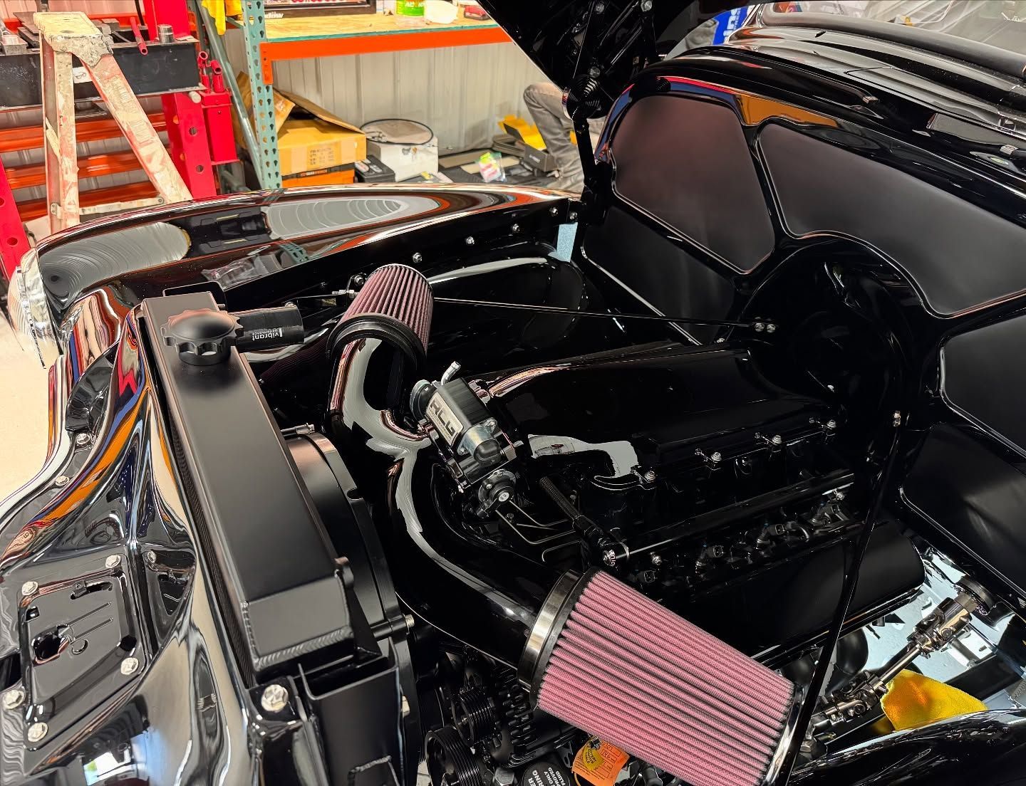 Open car hood showing a glossy black engine bay with pink air filters in a workshop.