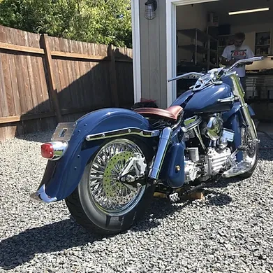 Blue motorcycle parked on gravel outside a garage with a person visible inside the doorway