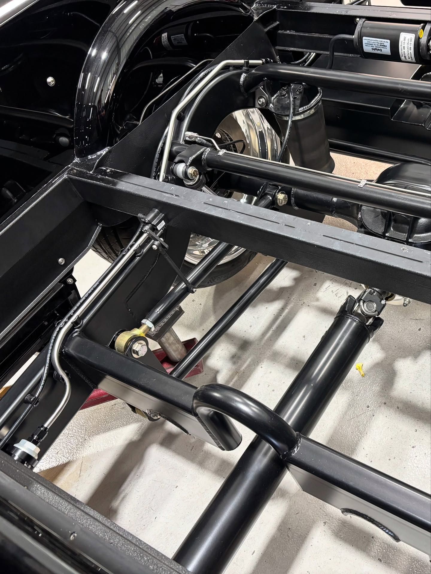 Close-up of a black metal vehicle chassis with axle, suspension, and wheels in a workshop.