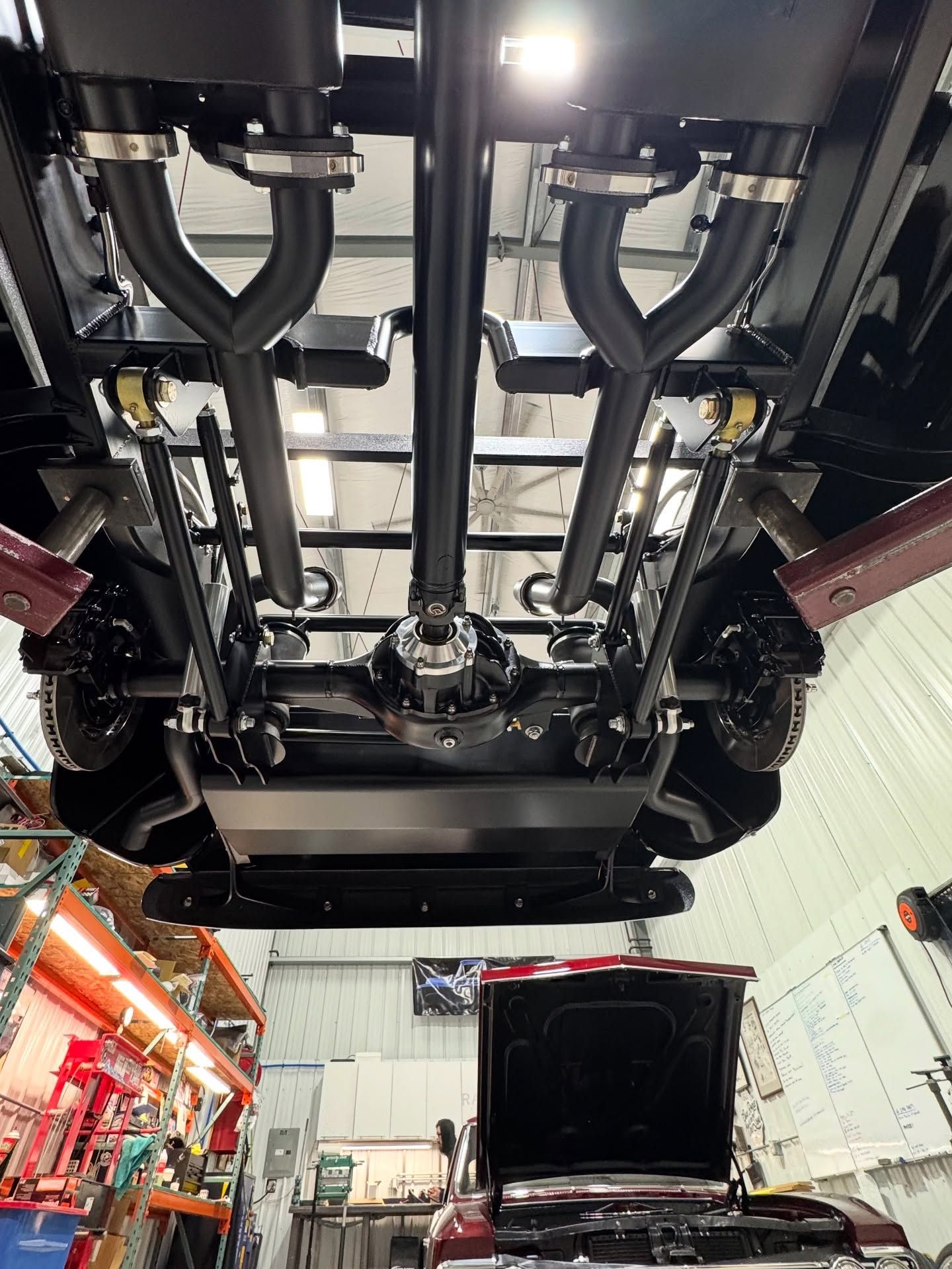 Undercarriage of a vehicle on a lift in a garage, with another car’s hood open below.