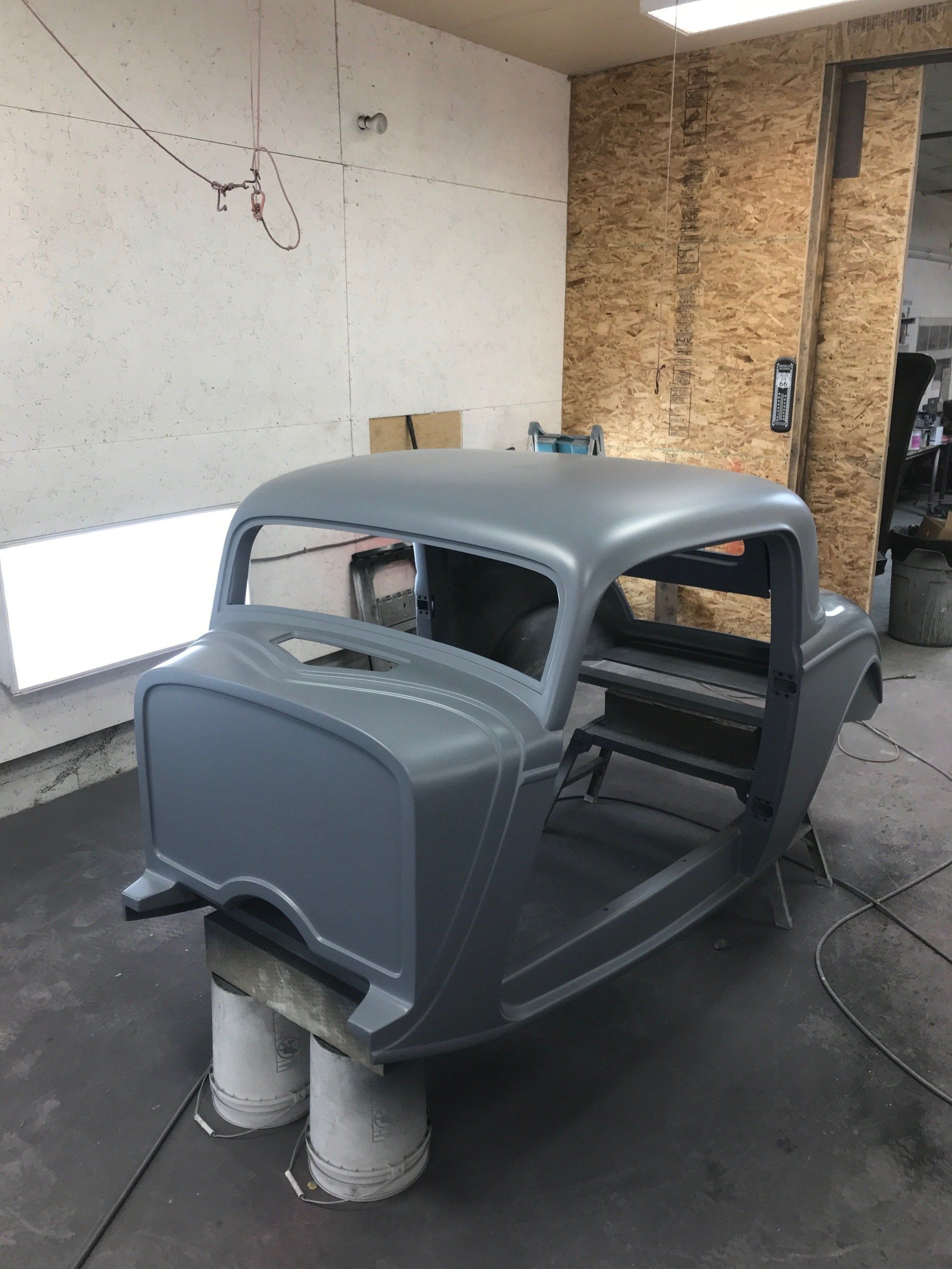 Gray vintage car body shell mounted on supports in a workshop, with unfinished walls and tools nearby