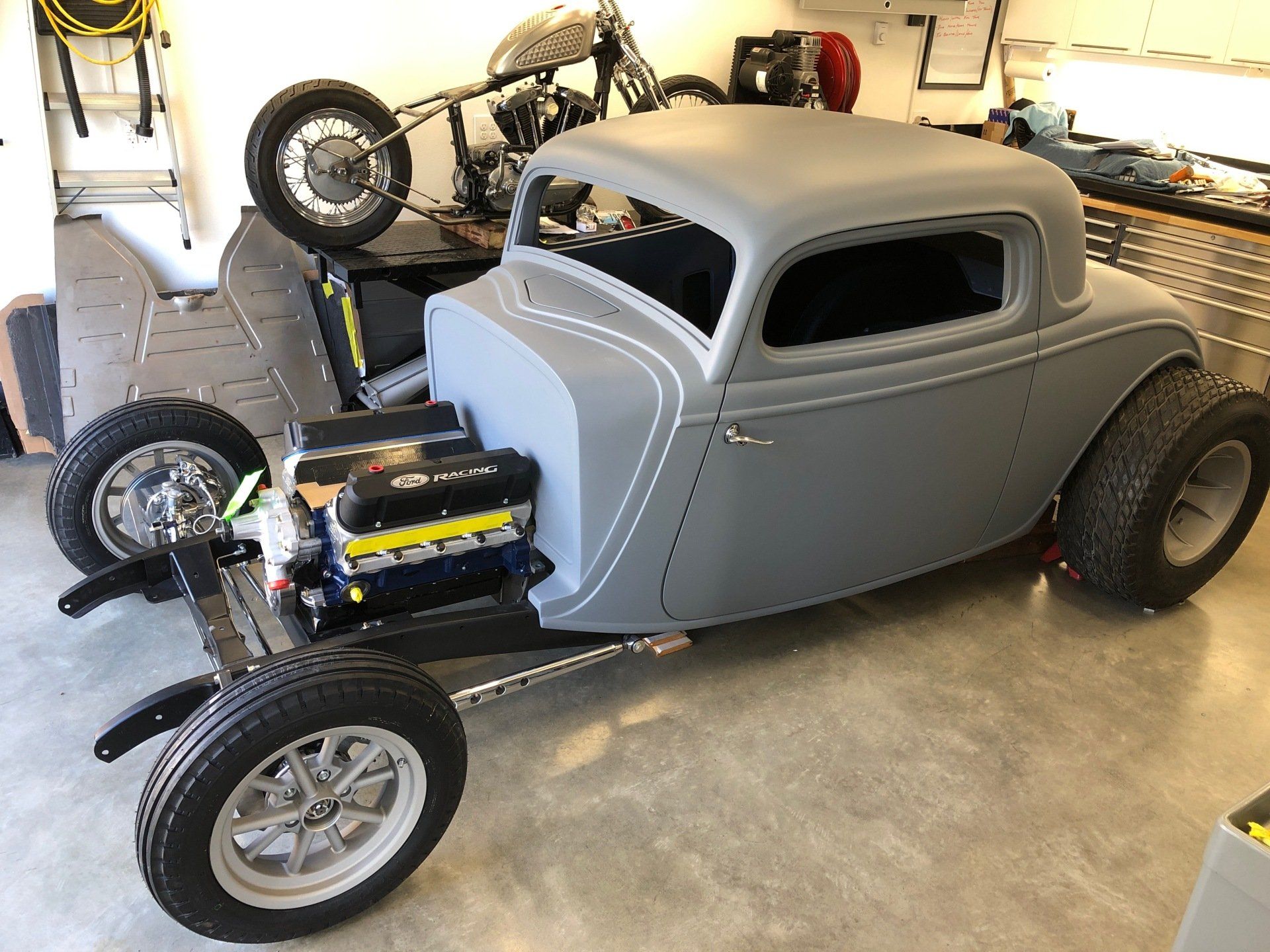 Gray hot rod coupe in a garage, with exposed engine and a motorcycle mounted on the roof.