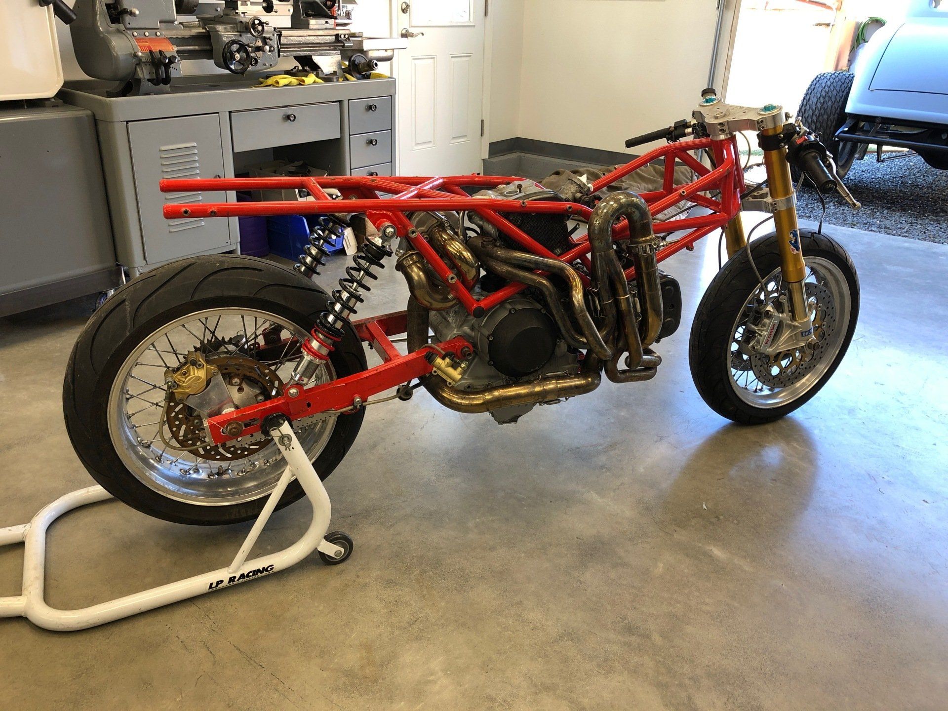 Red custom motorcycle frame in a garage, with exposed engine and front wheel on a stand on a concrete floor