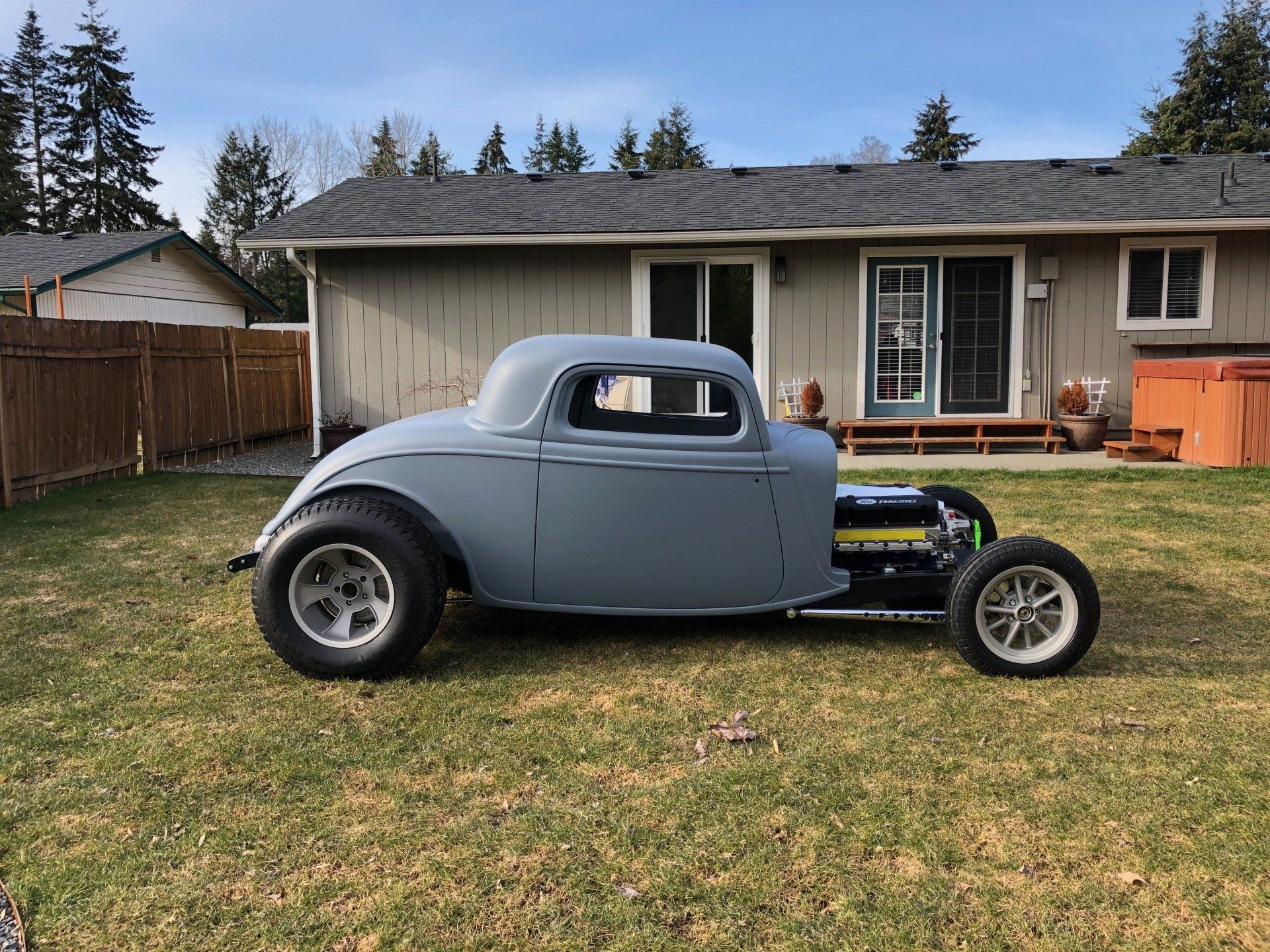 Gray classic hot rod car parked on a lawn in front of a suburban house.