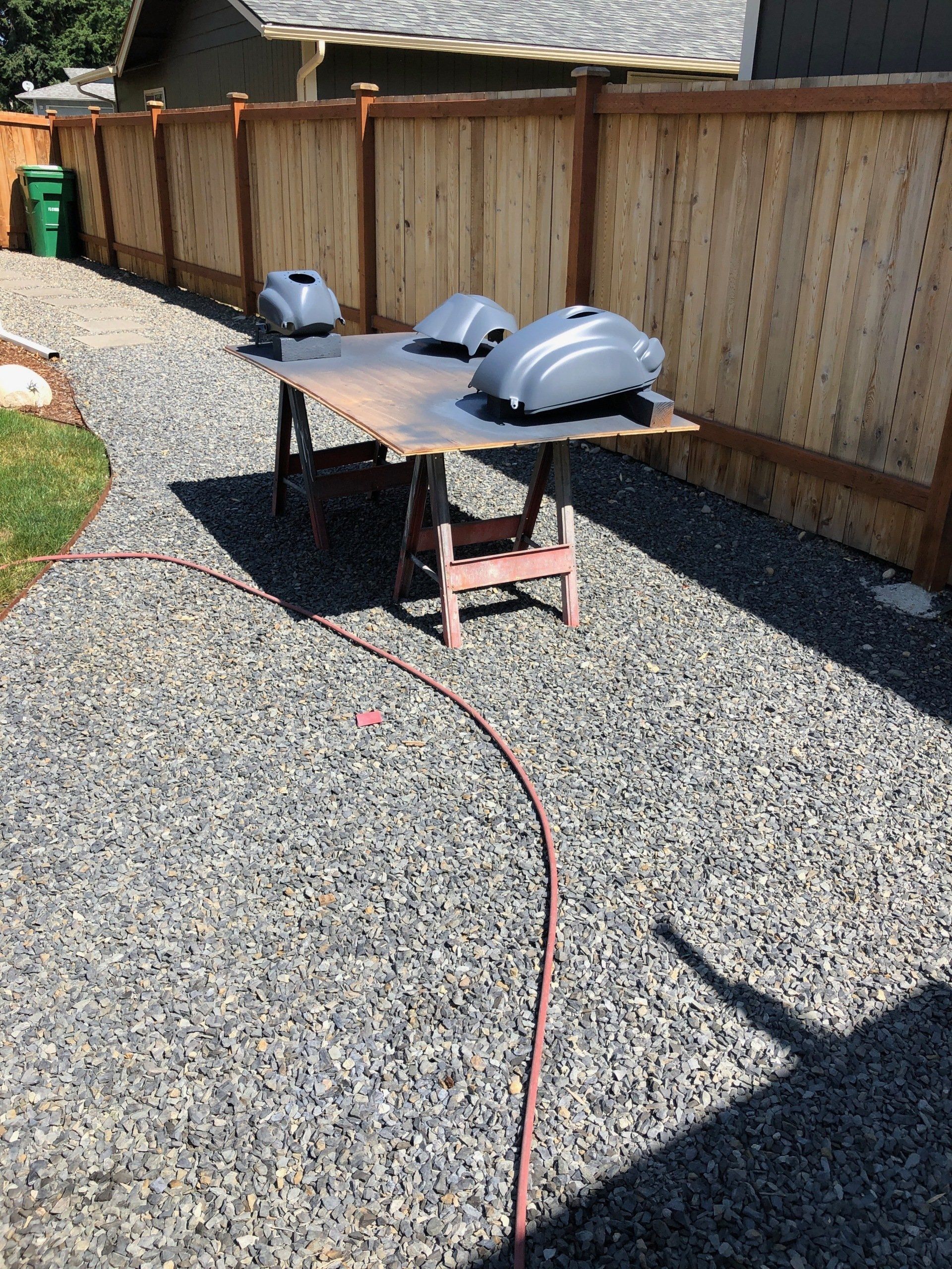 Outdoor workbench on gravel with two gray car hoods and a red hose beside a wooden fence.